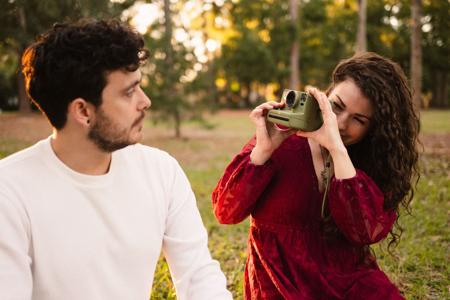 A couple in burgundy and white outfits share romantic moments during a golden hour photoshoot in a park setting.