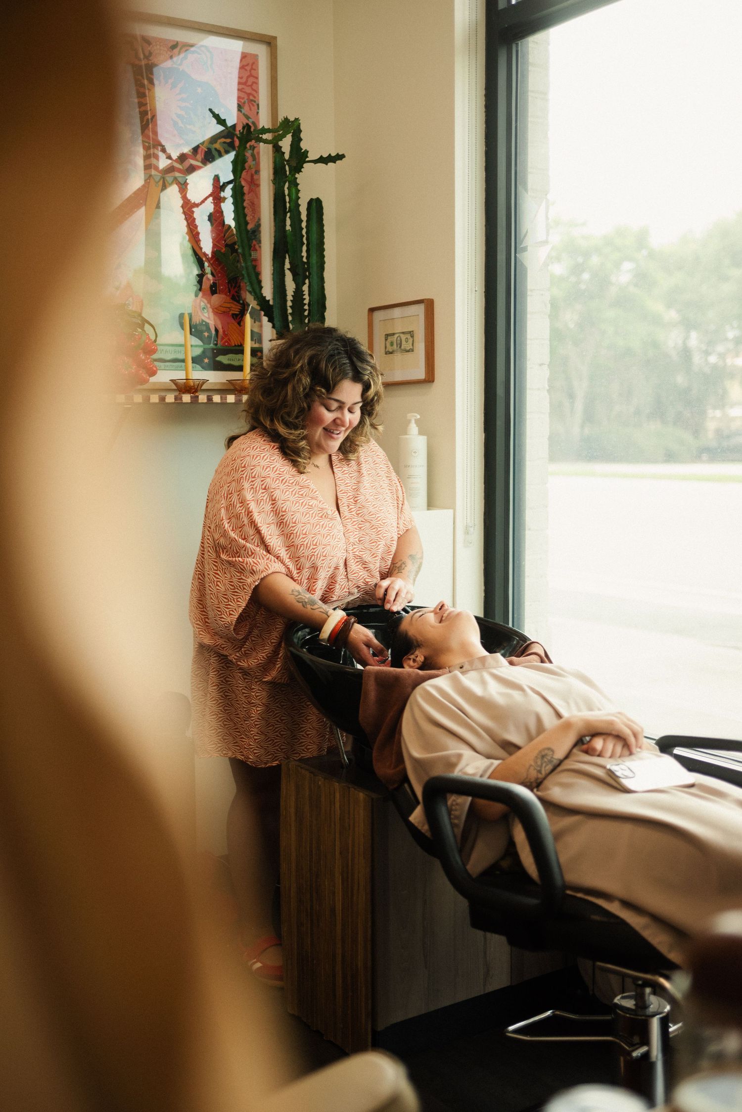 Sunlit salon scene showing a stylist working near large windows with plants.