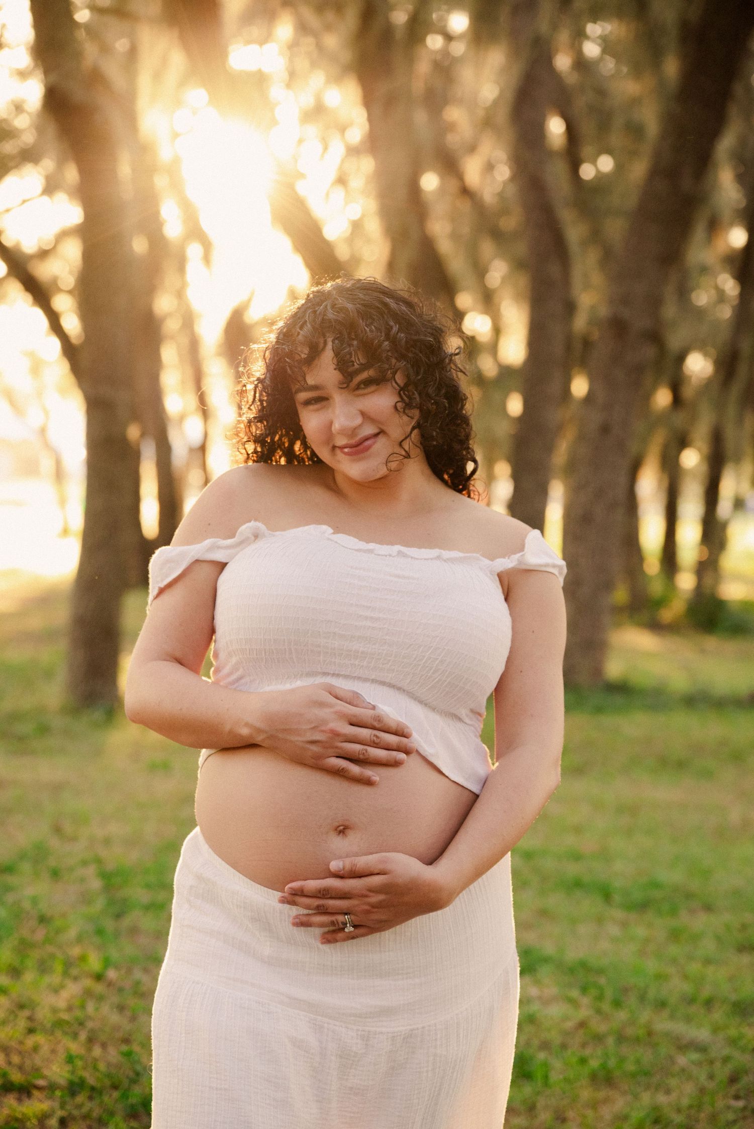 A maternity photoshoot in a white dress captures tender moments in golden sunlight.