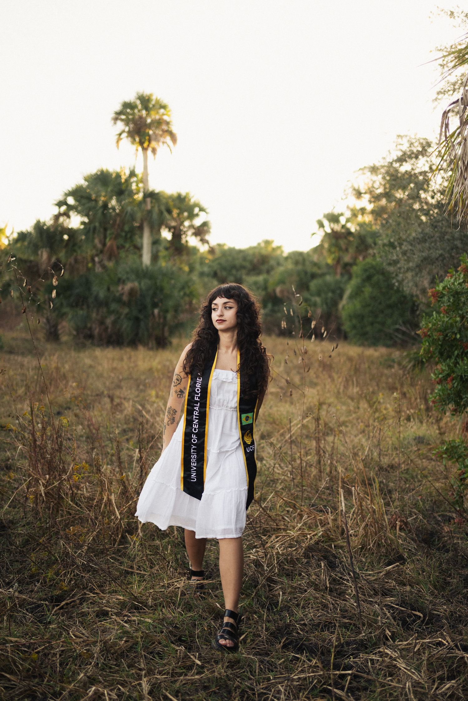 Graduate in white dress walks through tall grass during sunset graduation photoshoot.