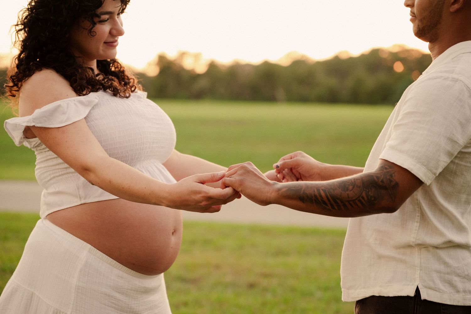 A couple holds hands in a sunlit field during a maternity photoshoot.