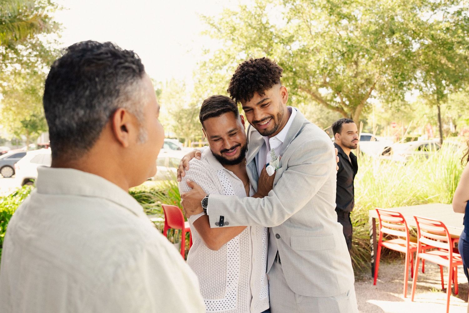 Wedding guests in light colored attire share joyful moments outdoors under trees.