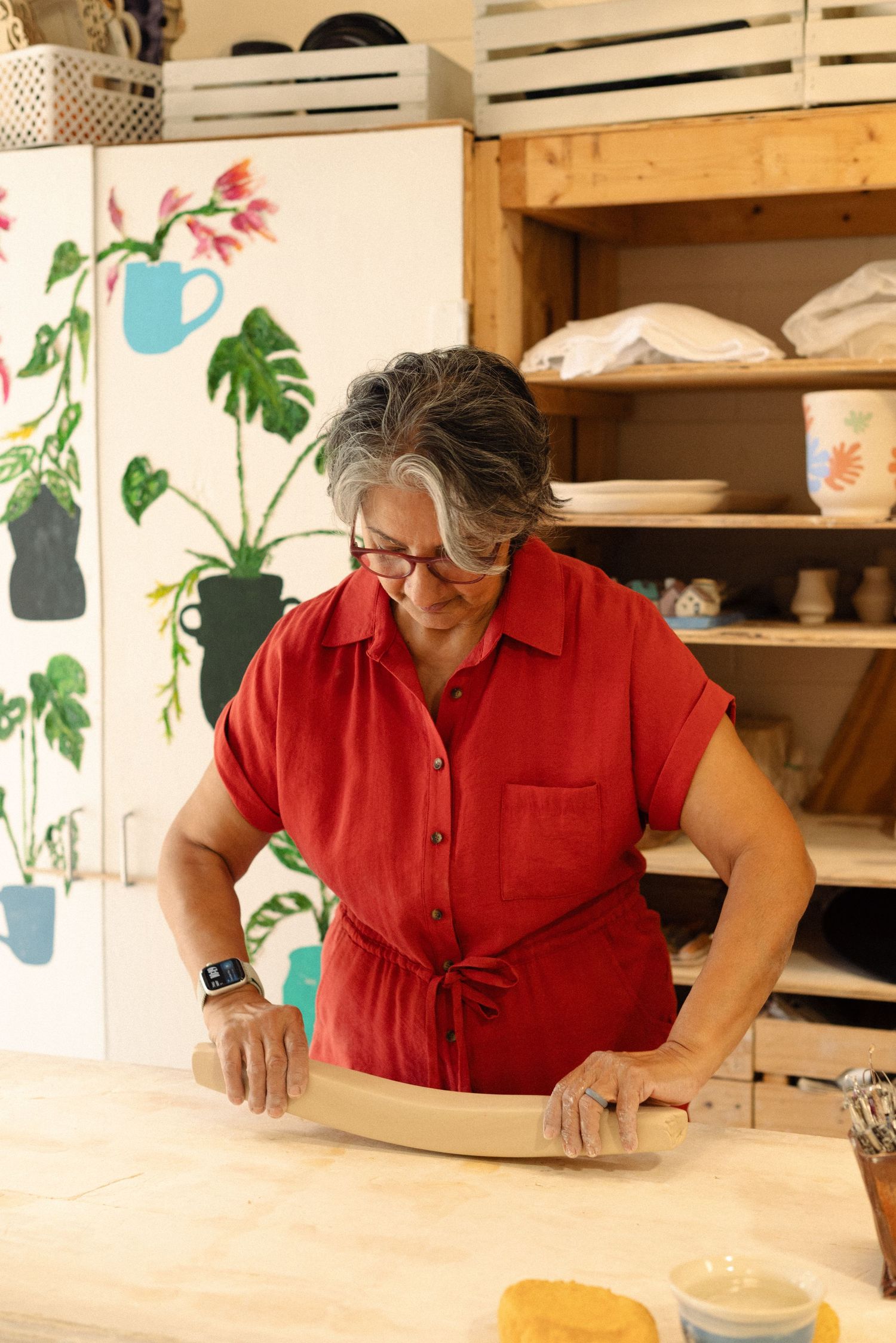 Artist rolling clay on a wooden work table in a studio with painted wall murals.