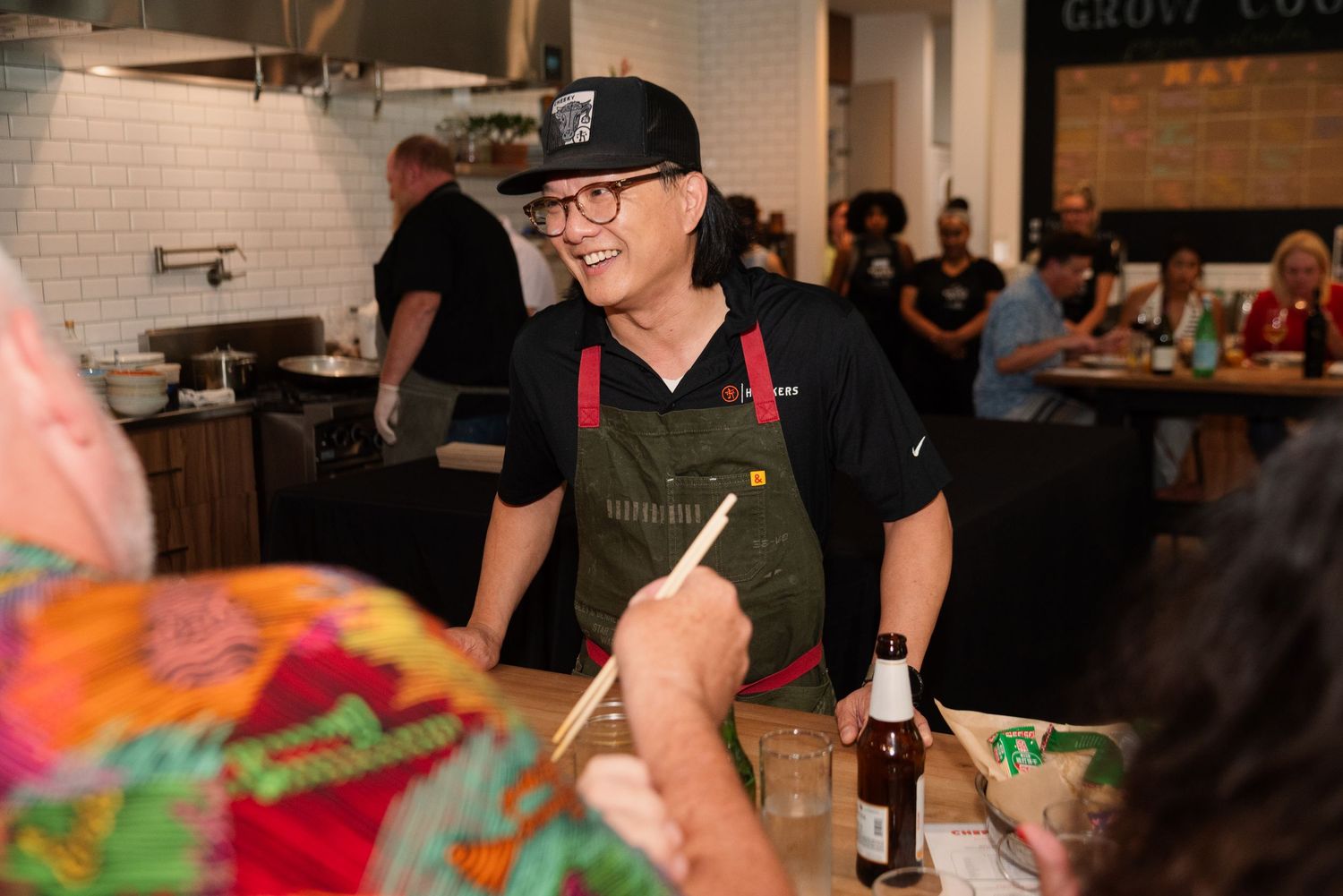 Restaurant staff member in apron and cap serving guests at counter with white tiled kitchen backdrop.