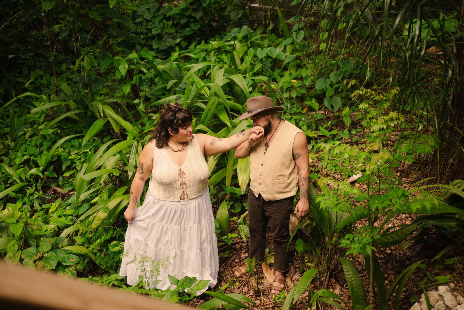 A couple walks together through lush green foliage in vintage-style clothing.