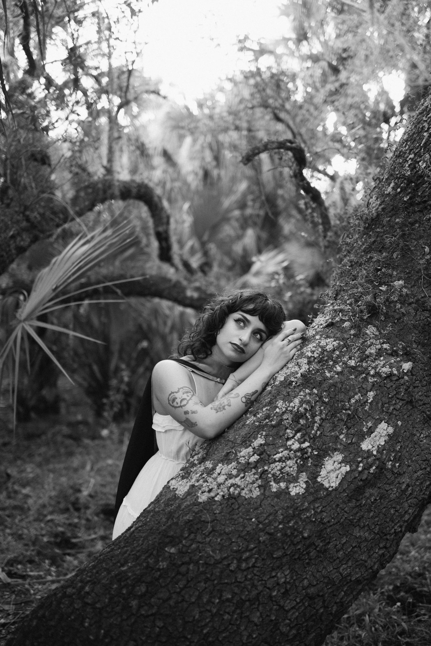 Black and white portrait of someone resting against a mossy tree in a dreamy forest setting.