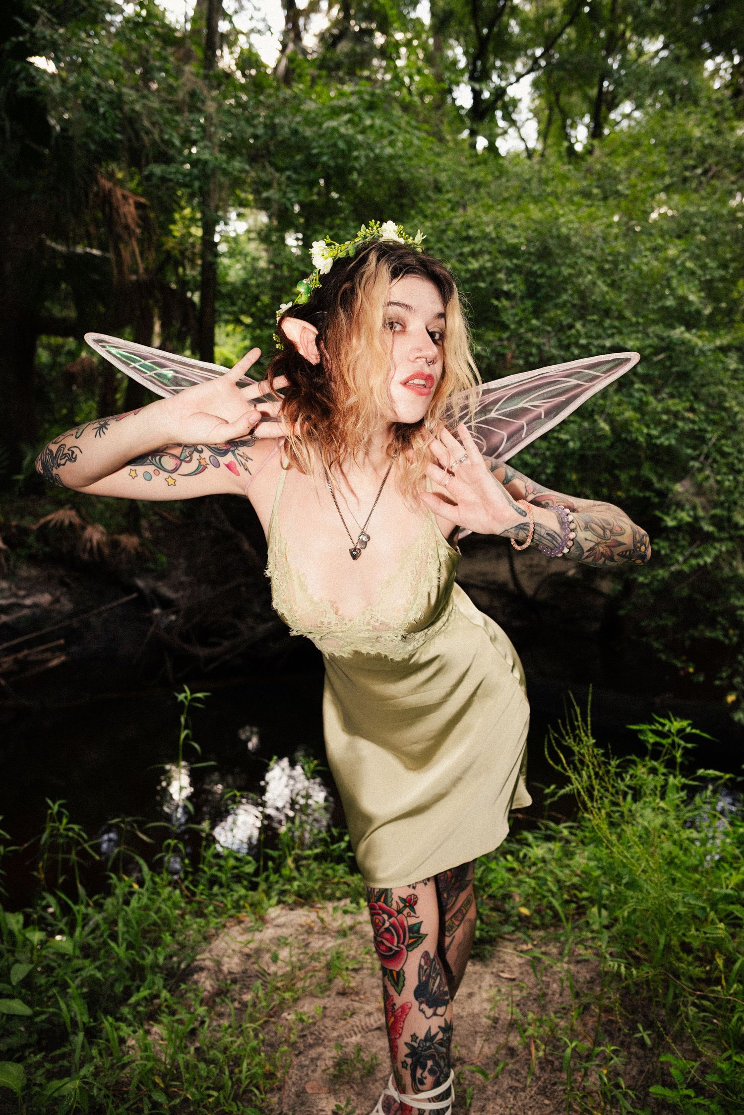 Whimsical fairy with tattoos and translucent wings poses playfully in a forest setting.