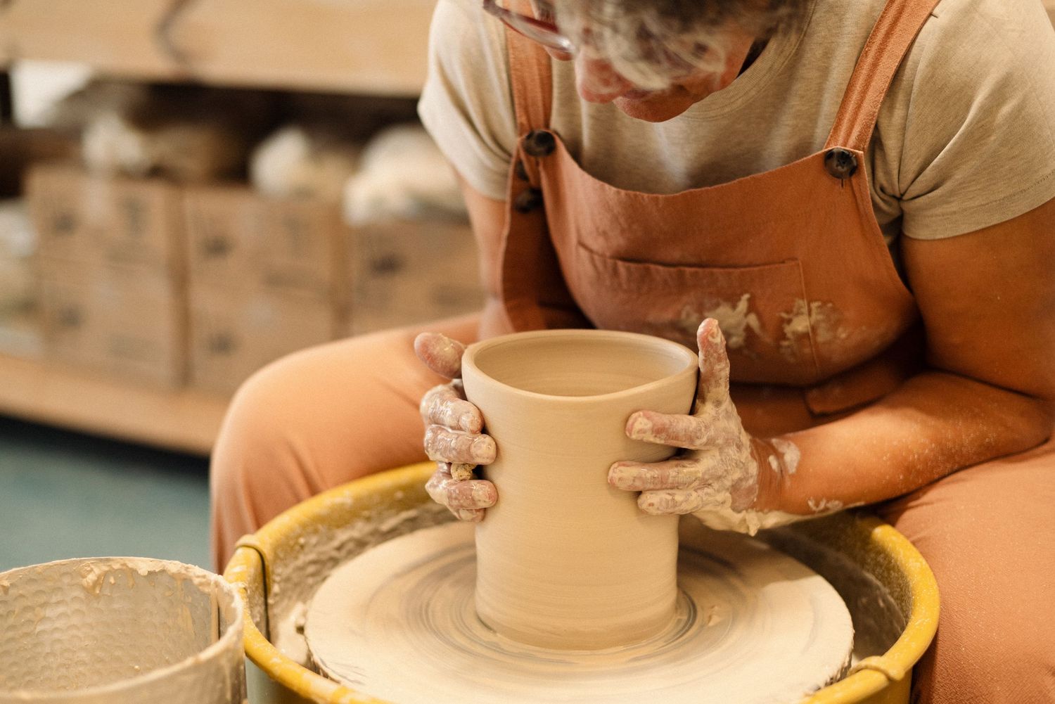 A potter shapes wet clay on a pottery wheel while wearing a brown apron in a ceramics studio.
