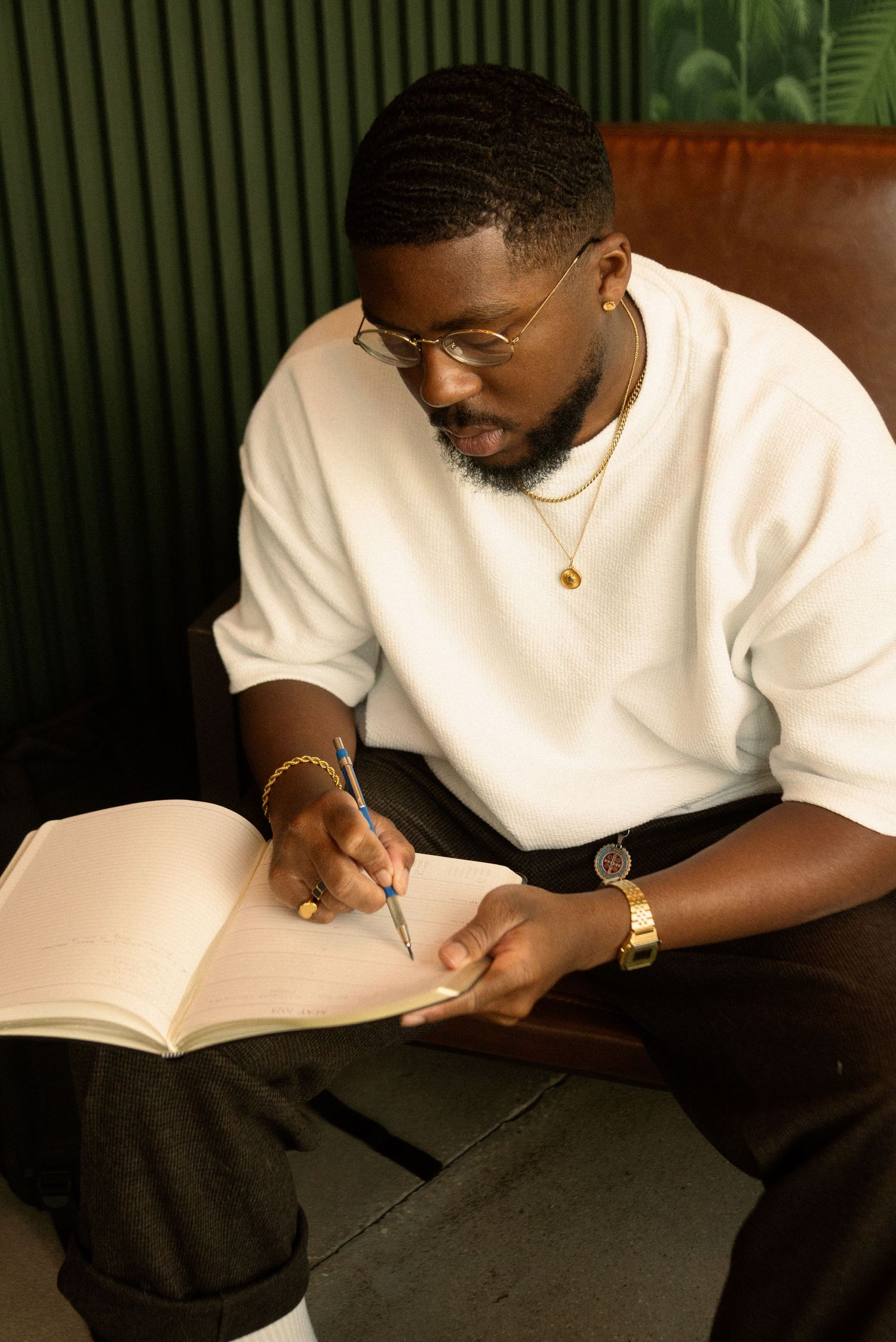 Person writing in notebook while seated in brown leather chair with green plants in background.