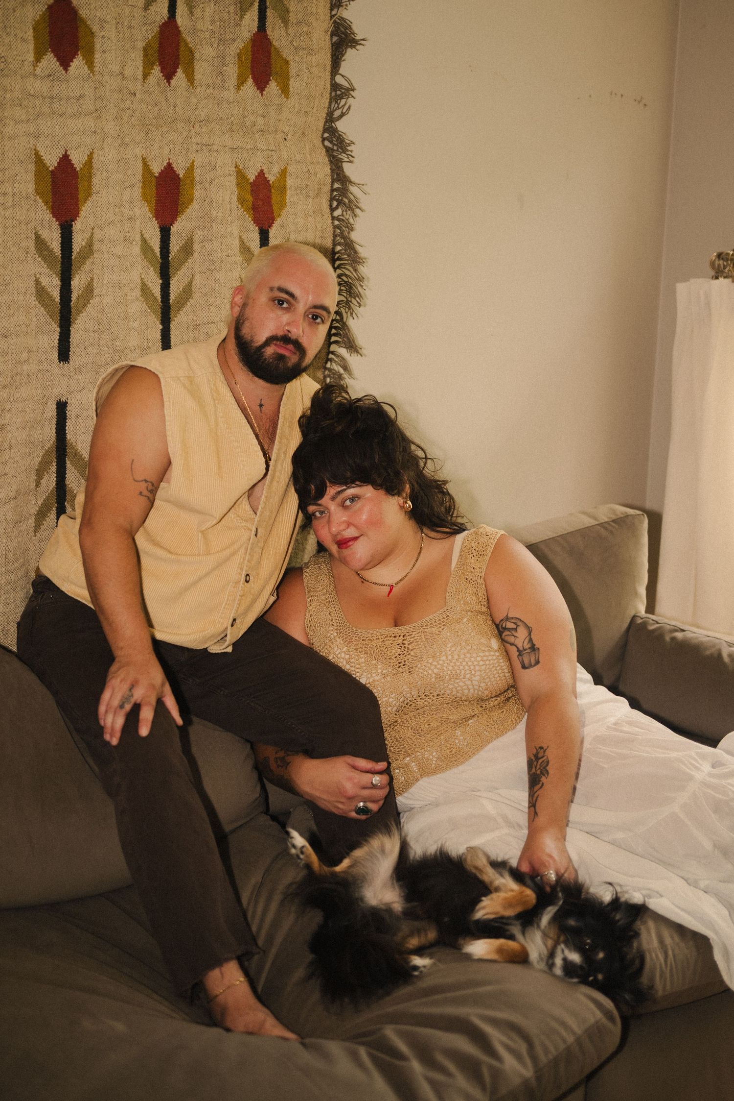 A couple in beige outfits sits casually on a couch with a black pet nearby.
