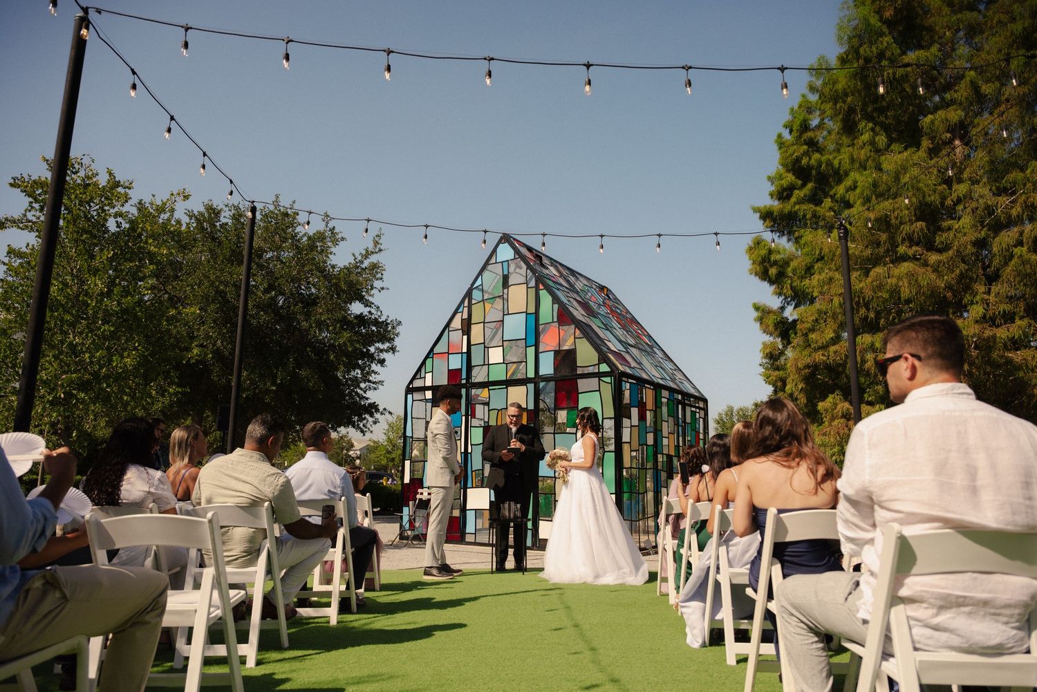 String lights hang over an outdoor wedding venue with white chairs set up on green grass.