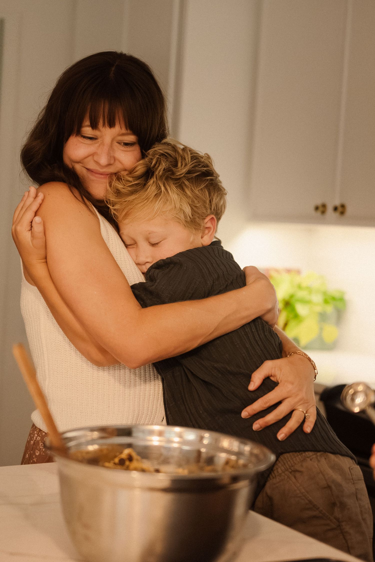 Two people share a warm embrace in a kitchen with jars and bowls visible on the counter.