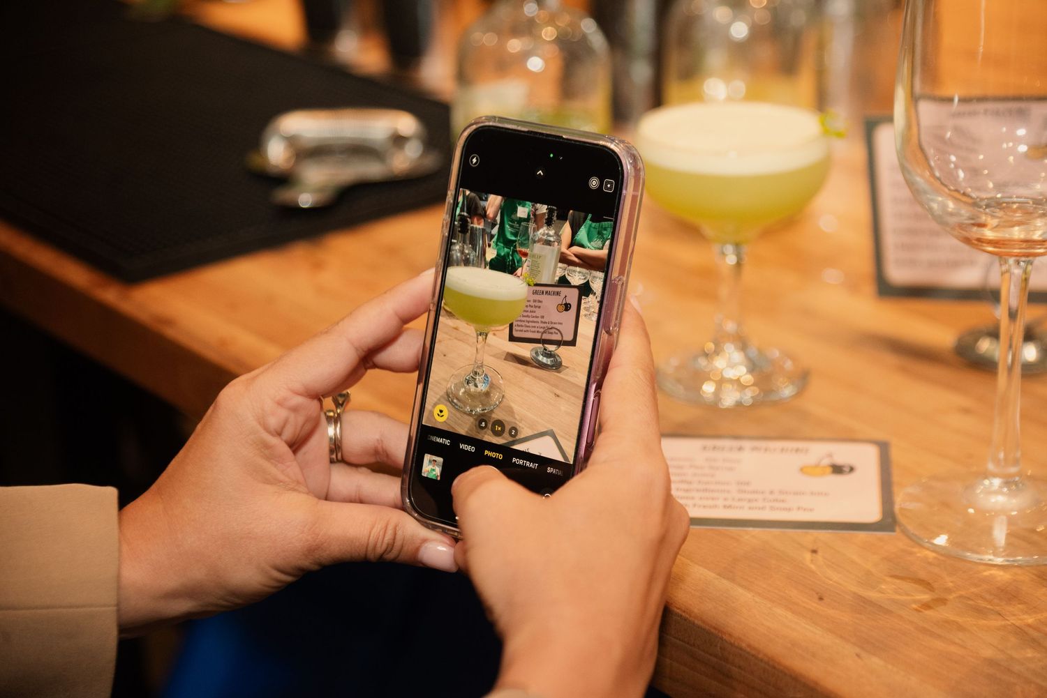 Cocktail glasses with garnishes sit on a wooden bar top while hands hold smartphones to take photos.