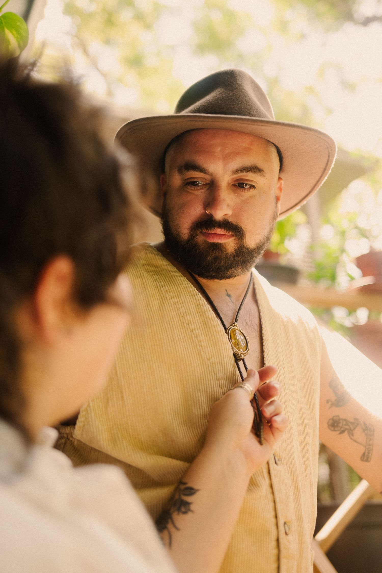 A person in a beige vest and hat poses during a sunlit outdoor photoshoot.