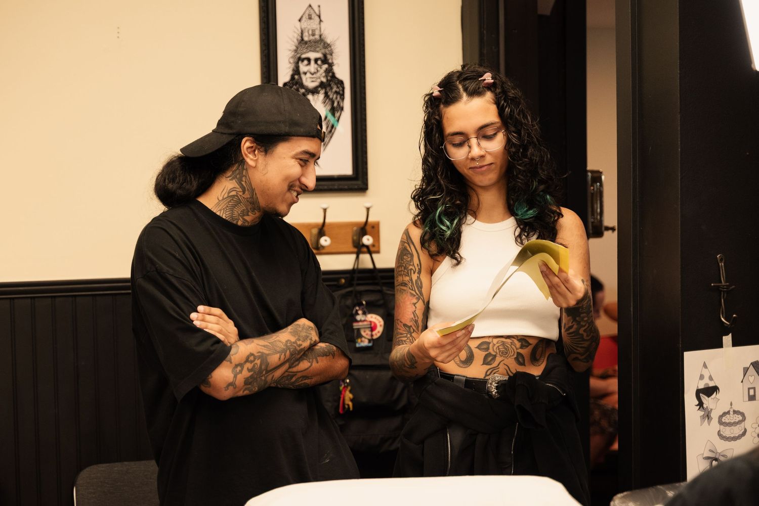 Artists collaborating in a tattoo studio with framed artwork on dark walls.