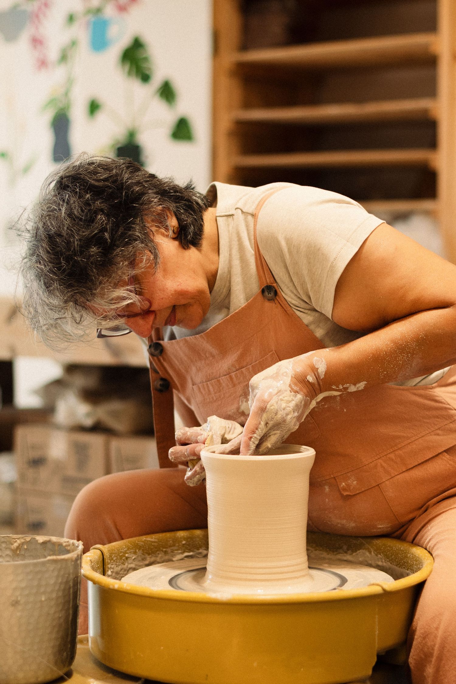 Potter shapes clay vessel on wheel in studio with painted botanical wall decor.