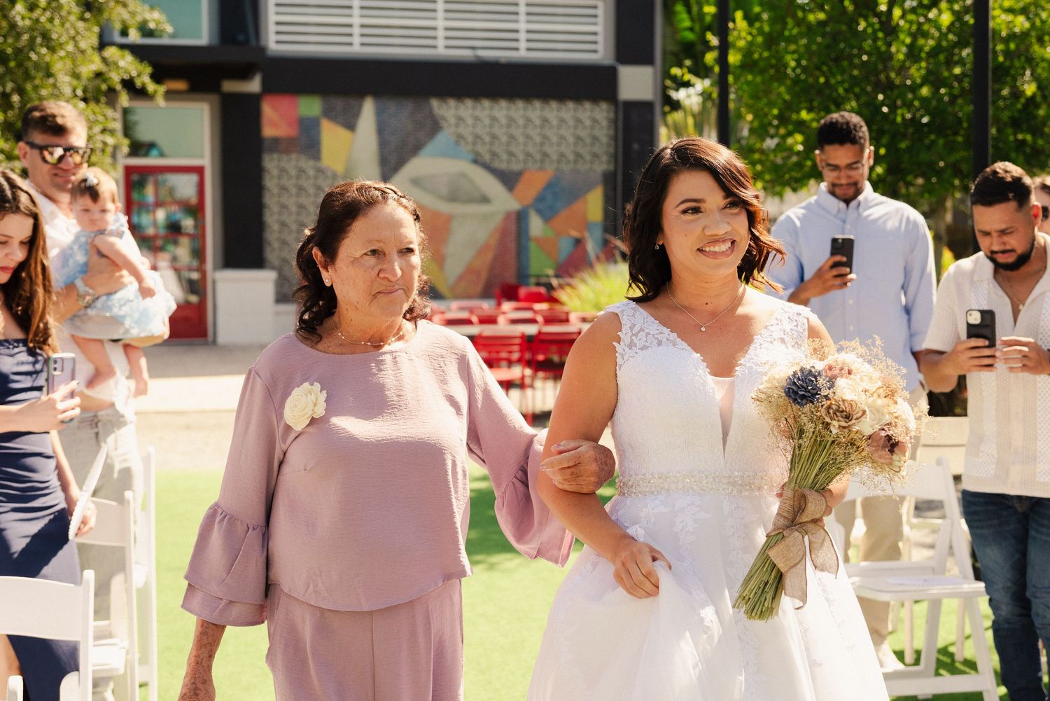 Wedding party members in pink and white attire gather outdoors during a sunny celebration.