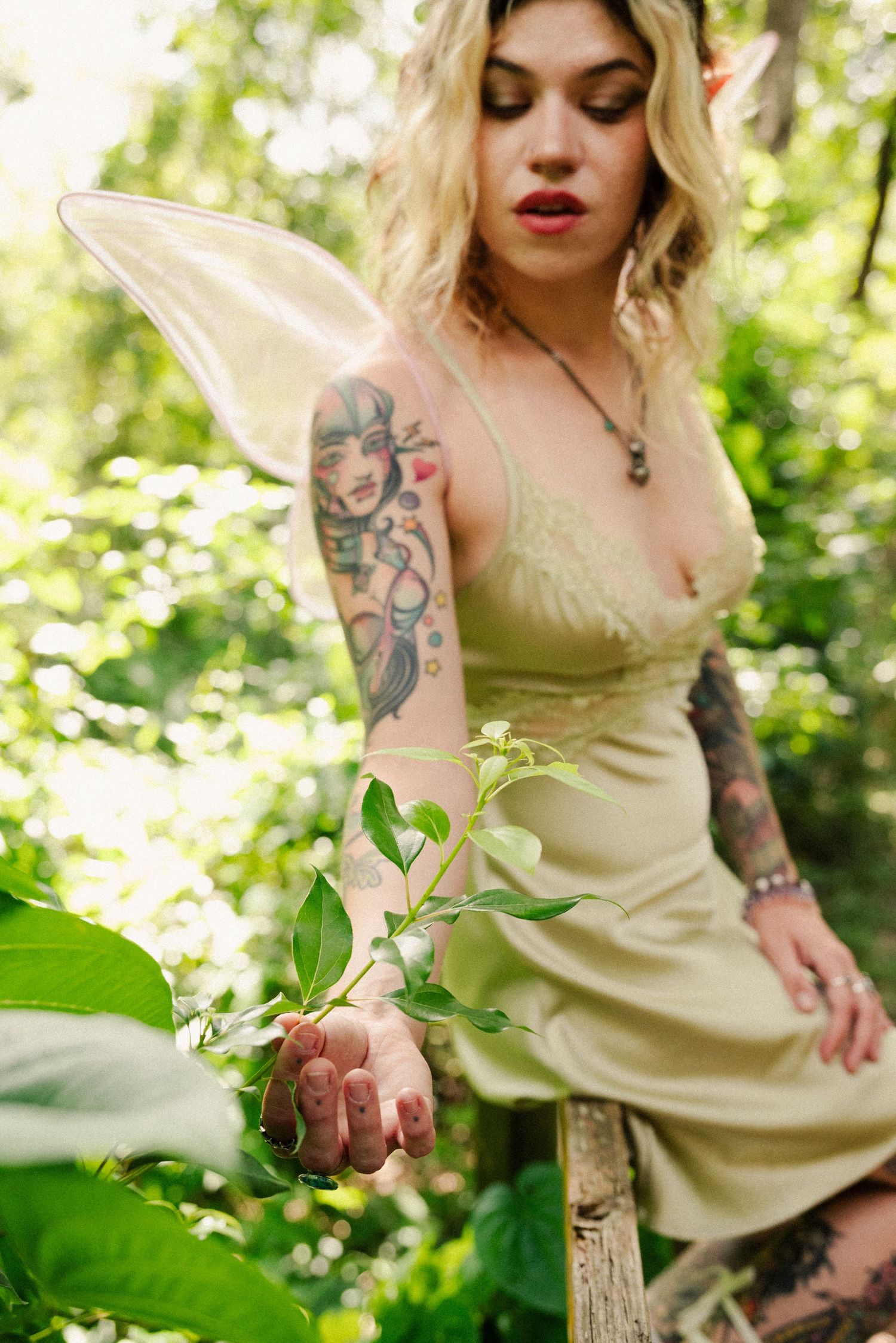 Tattooed fairy in cream silk dress poses among lush greenery wearing delicate wings.
