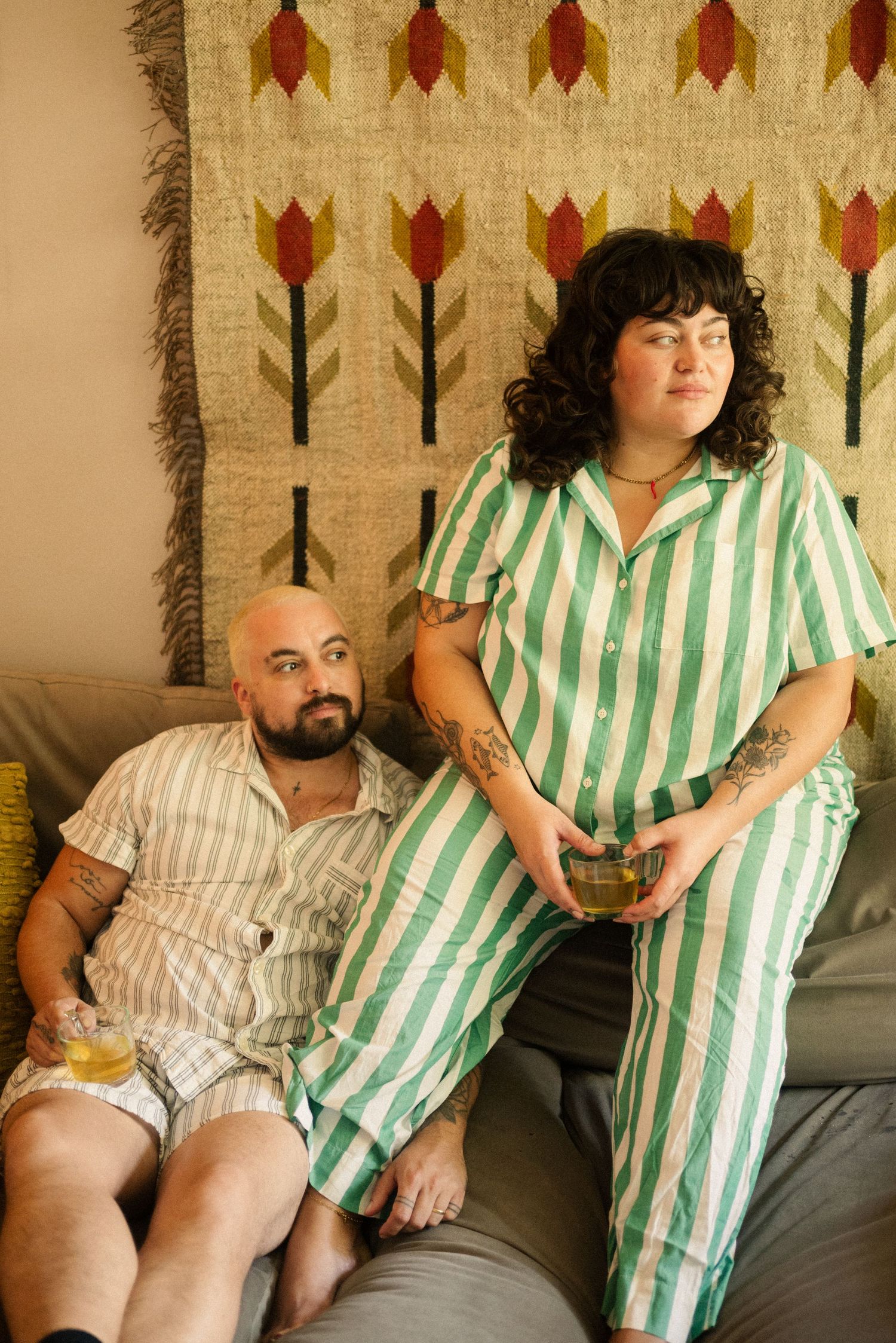 Couple in striped pajamas relaxing on couch with drinks against wall decorated with tulip pattern.