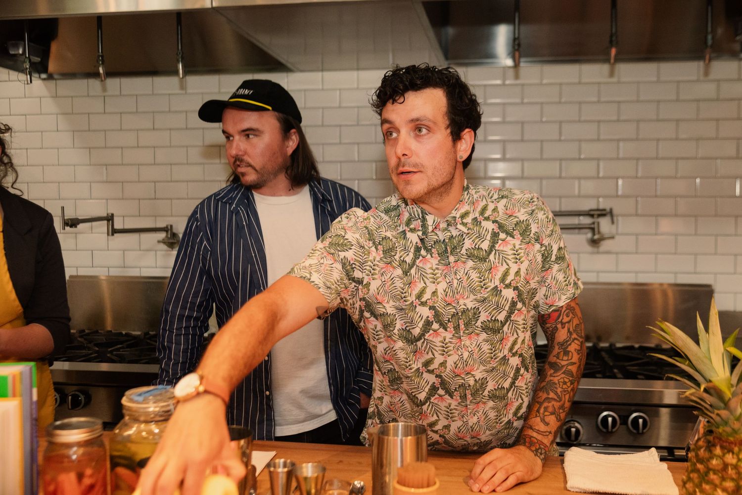 Participants in aprons gather around a cooking demonstration table in a modern kitchen with white subway tile walls.