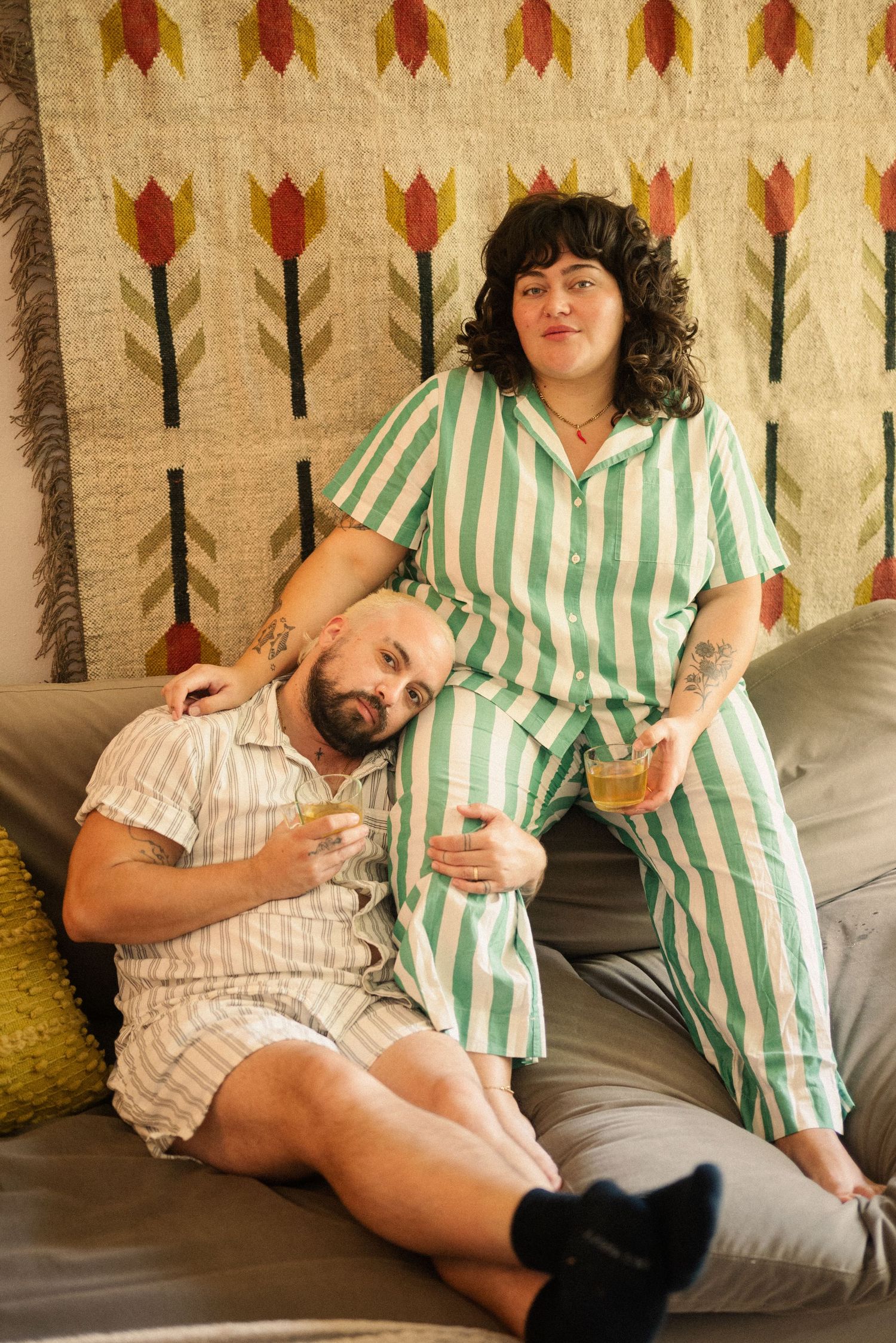 A couple in matching striped pajamas lounges together on a couch with decorative wall tapestry in background.