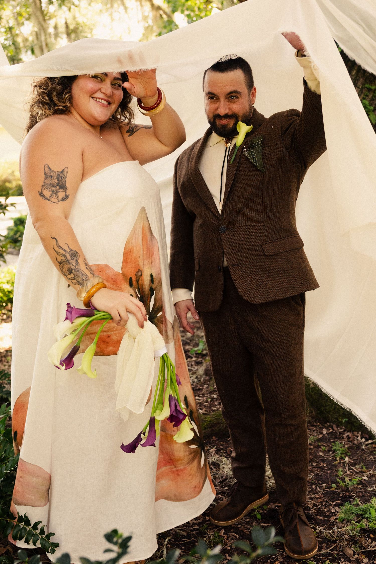 A wedding couple celebrates under white fabric draping, with the bride wearing a floral dress.