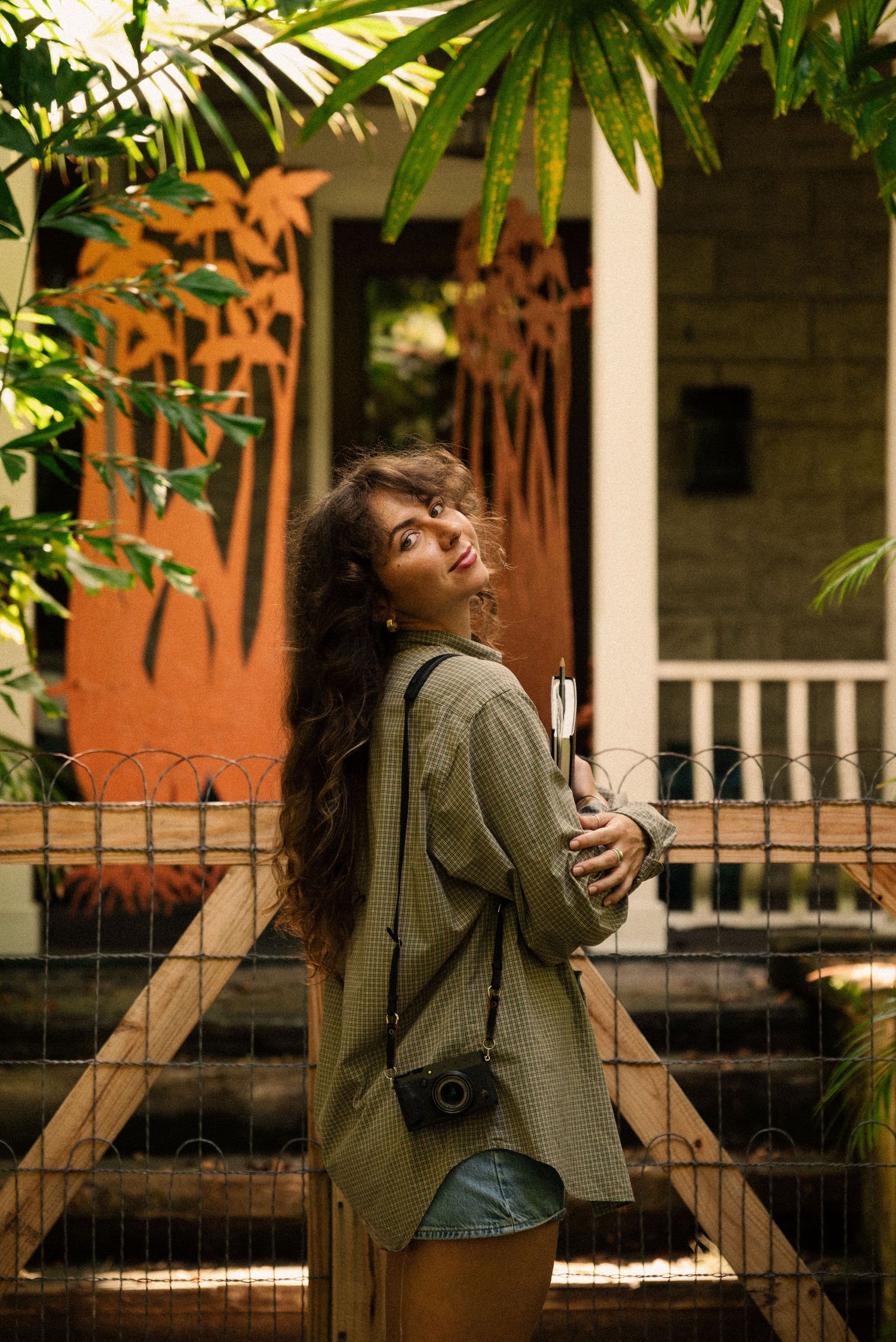 Figure in olive jacket leans back against a brick building with palm fronds overhead.