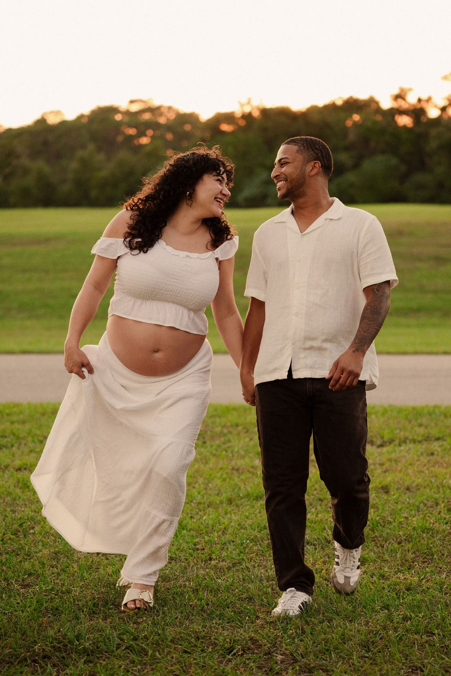 Playful maternity photos showcase couple dancing in grassy field at sunset.