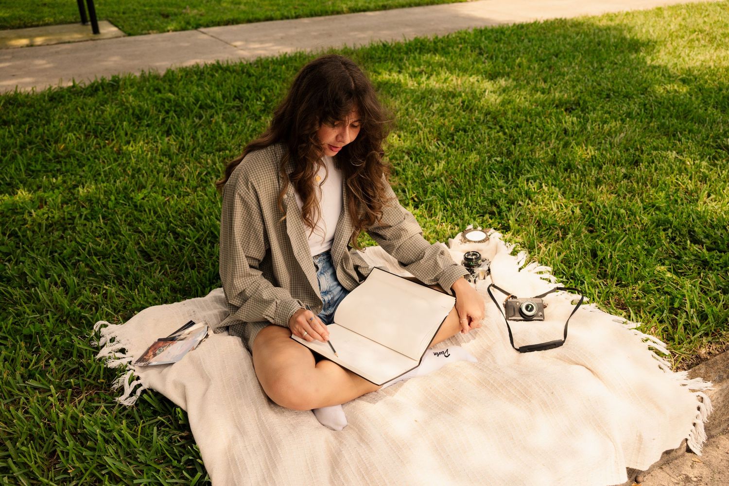 Person lounging on white blanket with notebook and camera in sunny garden setting.