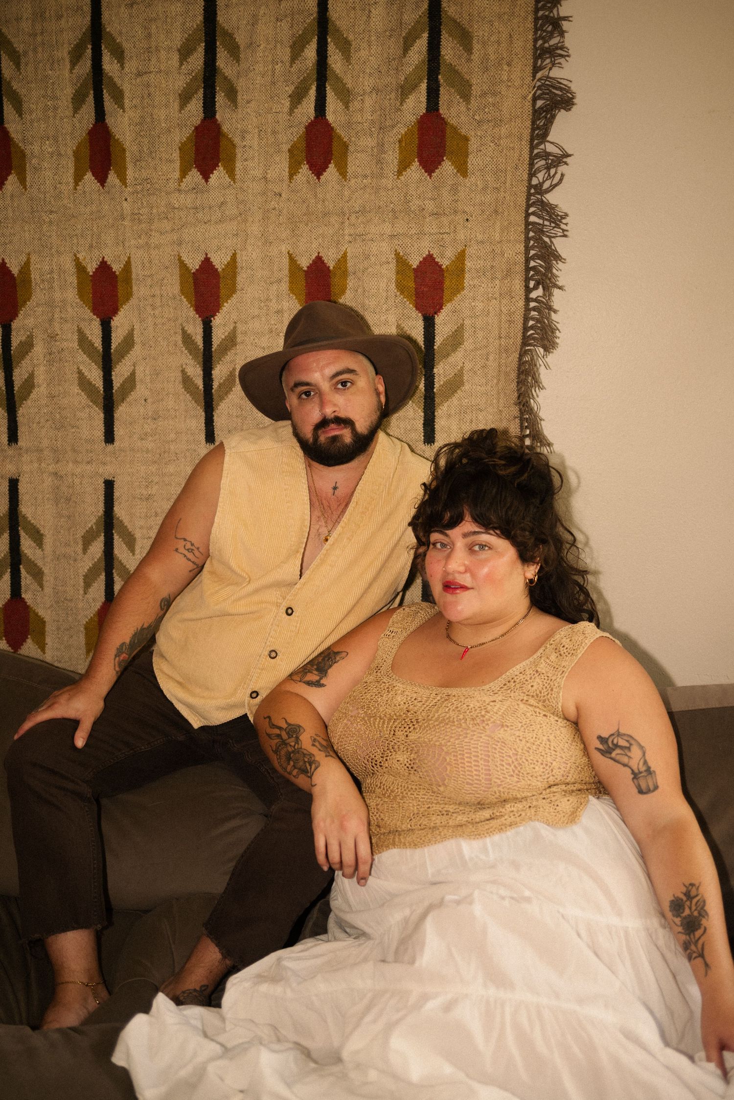 Couple in vintage-style clothing posing on couch against decorative wall hanging.