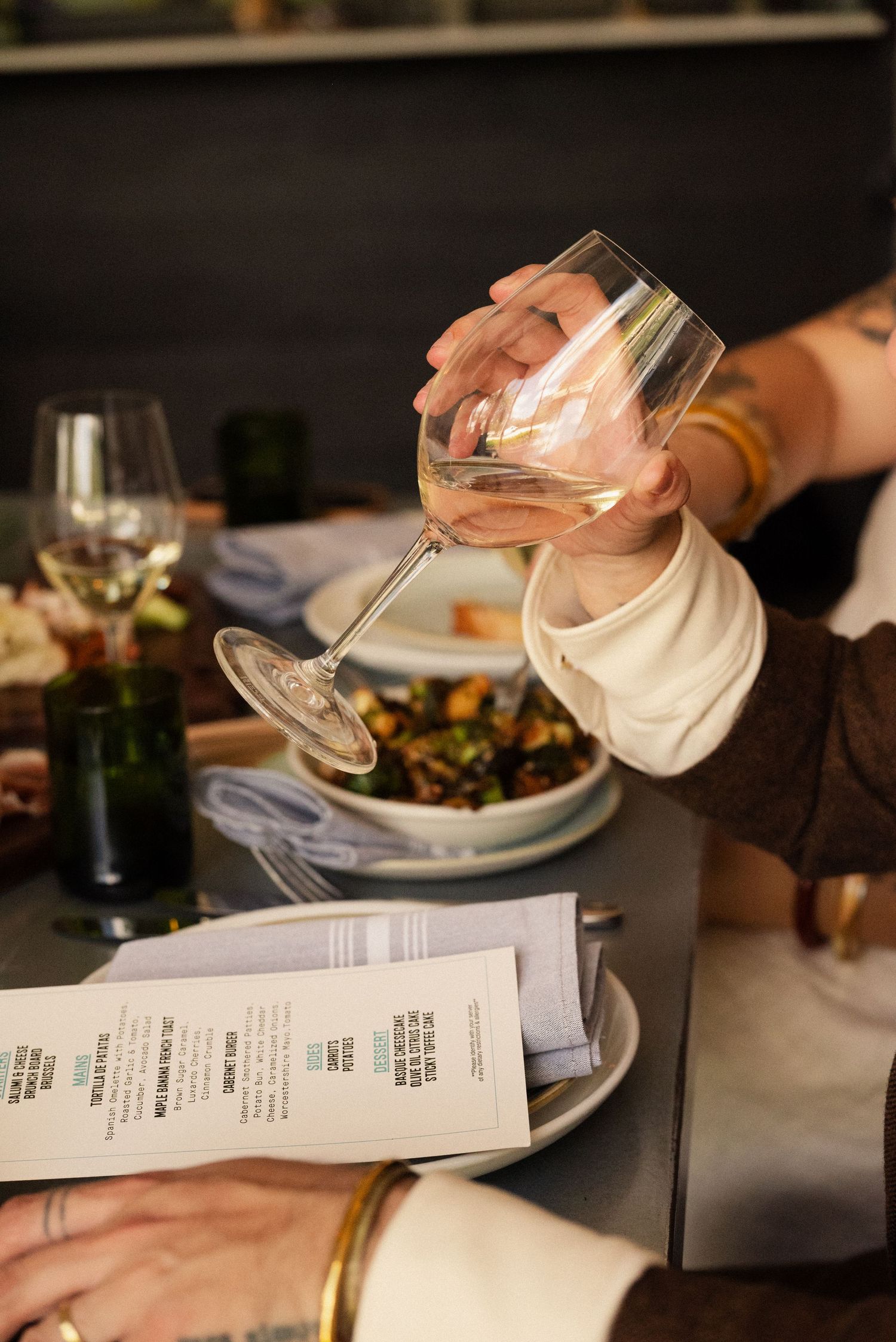 Hand holding a wine glass over a restaurant table with menu and place settings.
