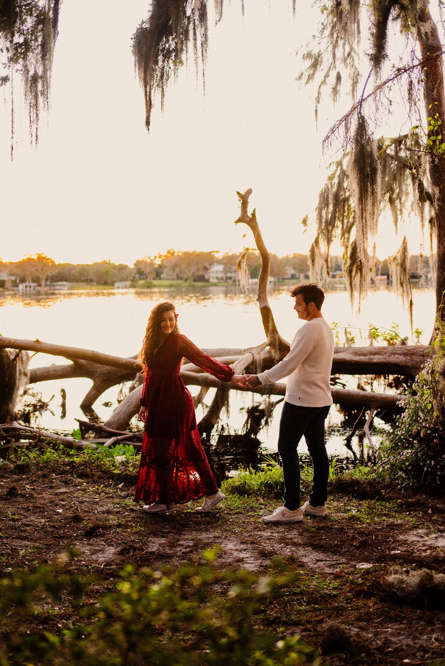 Couple sits by a lakeside at sunset under Spanish moss draped trees.
