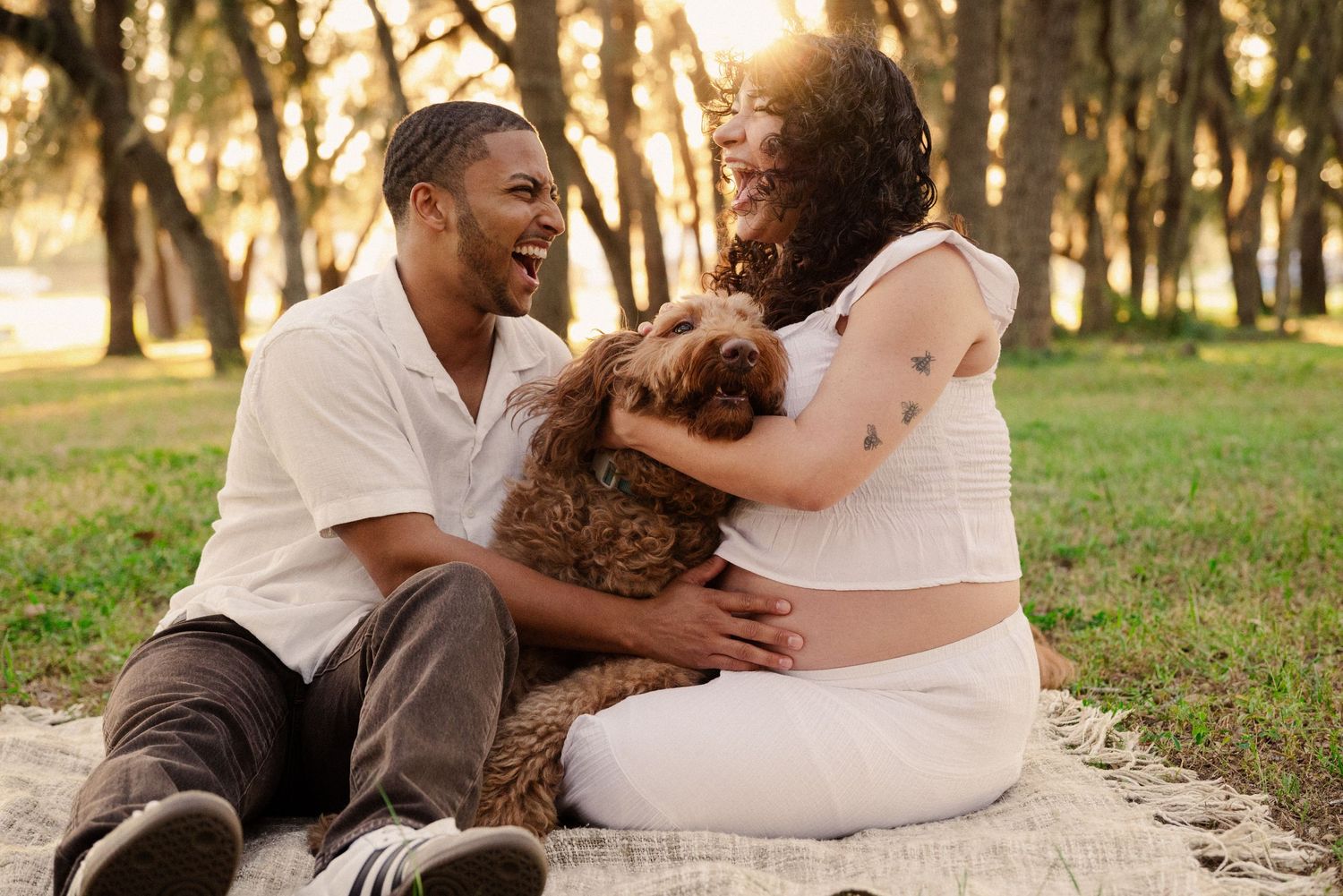 A loving couple and their dog pose for photos on a blanket in a sunlit park.