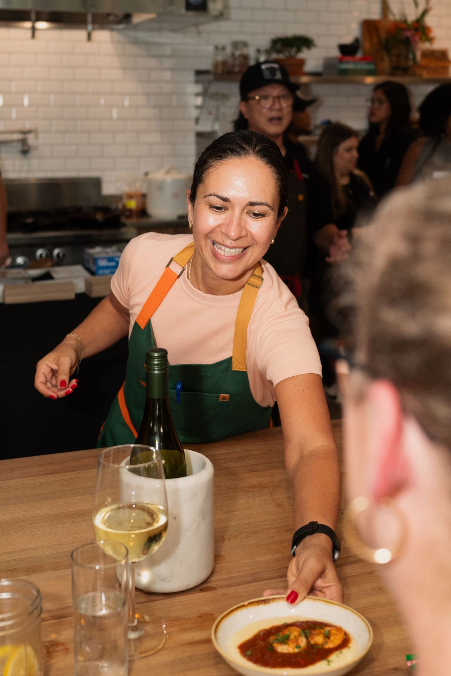 Server in green apron pouring wine at wooden counter in restaurant with white tiled kitchen.
