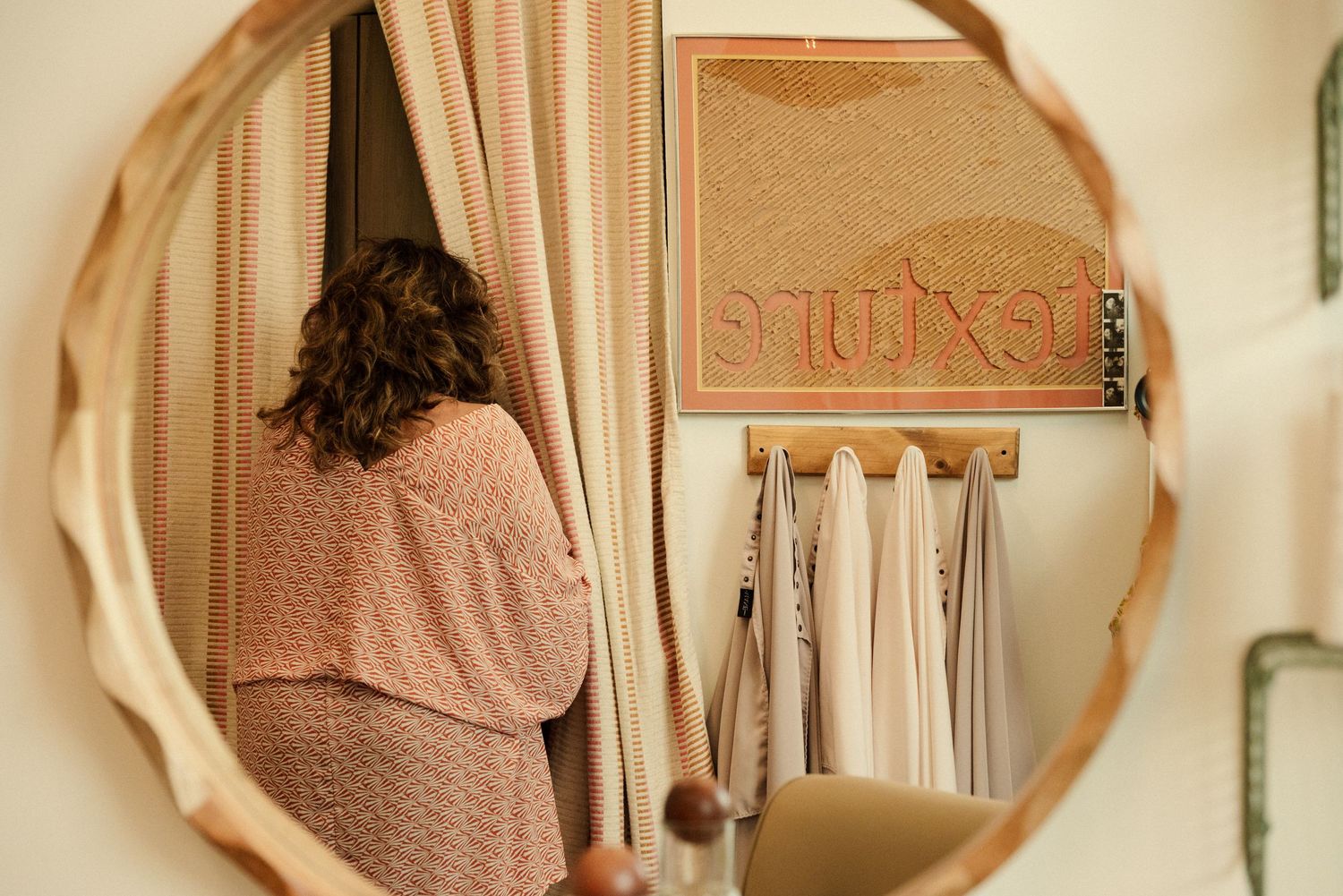 Round mirror reflects pink sweater and hanging clothes in a cozy bedroom setting.