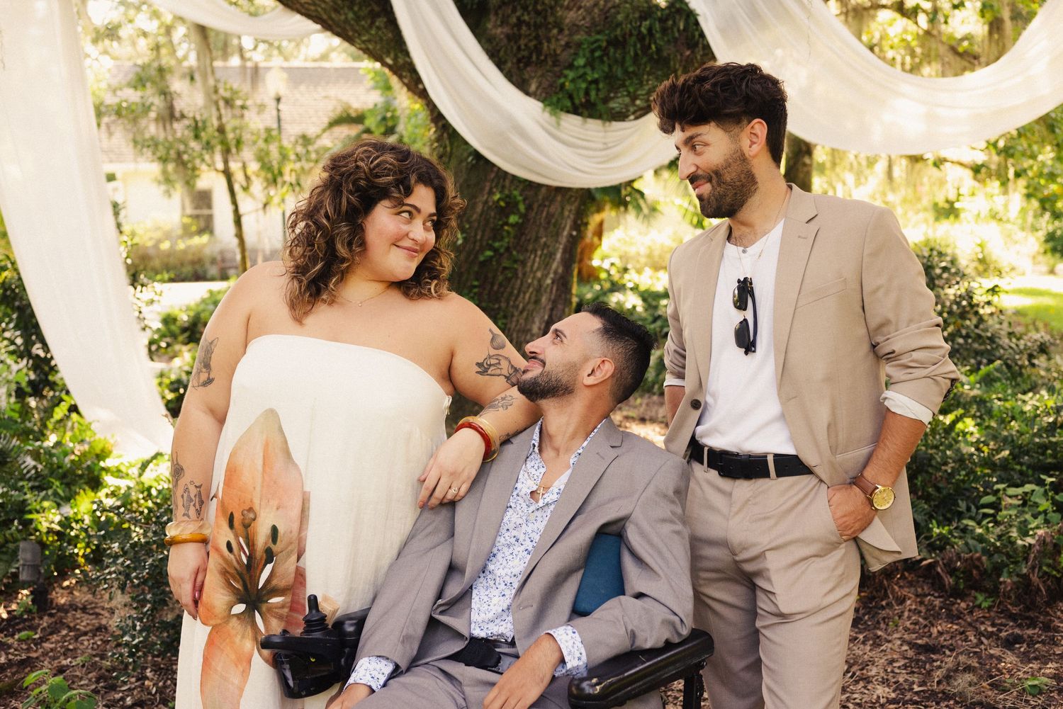 Wedding party poses casually outdoors under draped white fabric and trees.