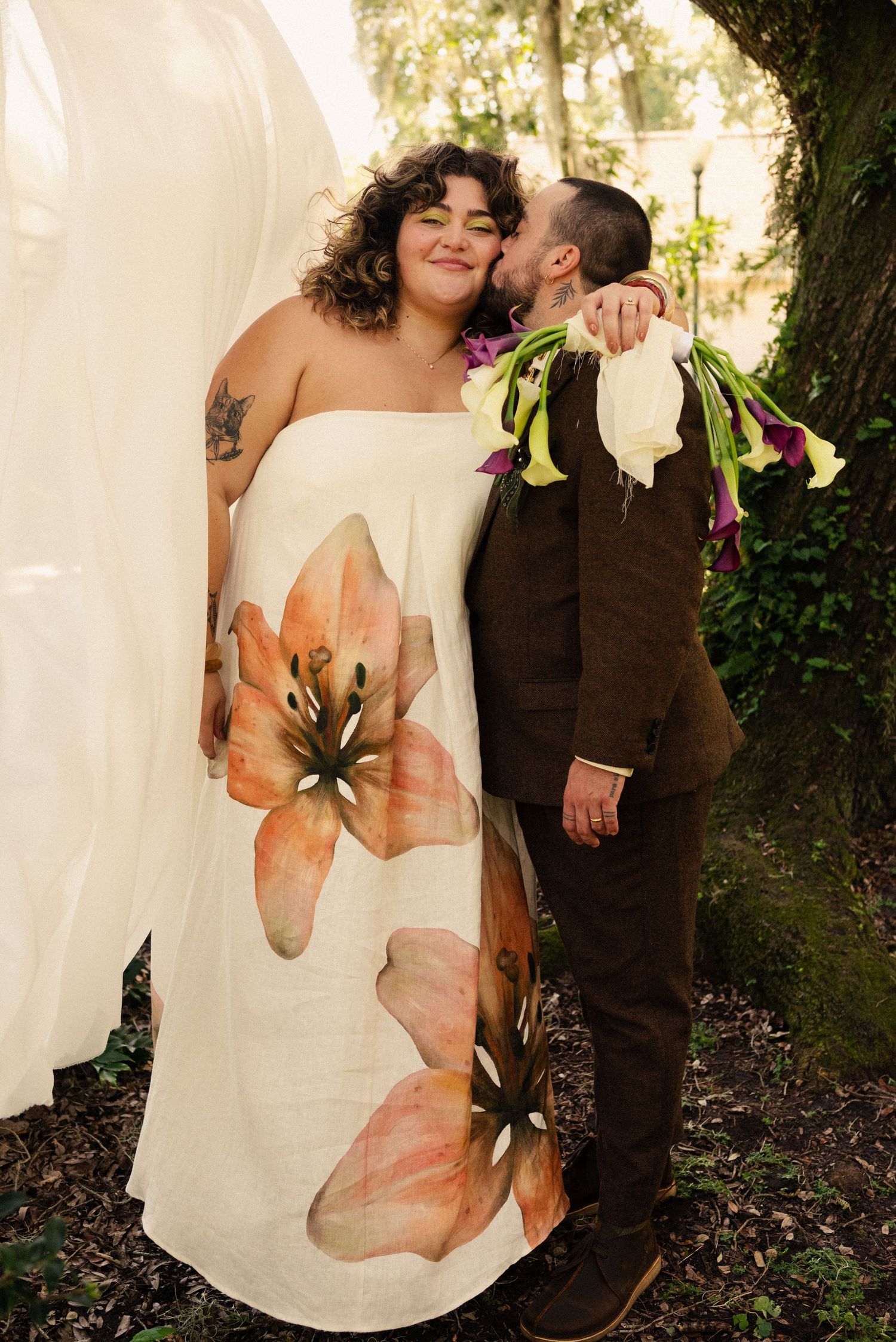 A couple in wedding attire stands together against a backdrop of trees, with a floral-printed white dress.