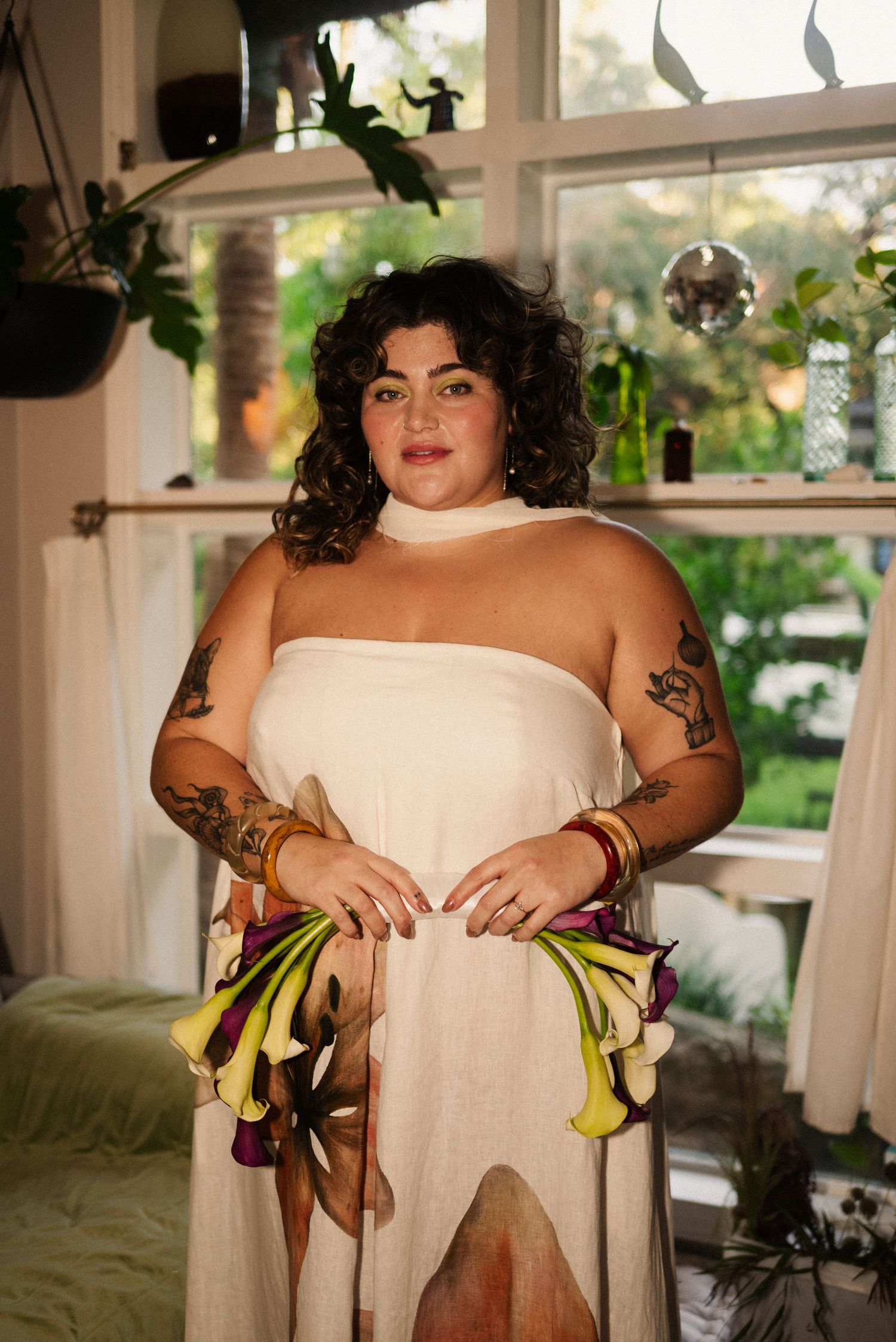 Person in white dress holds yellow calla lilies in a sunlit room with indoor plants.