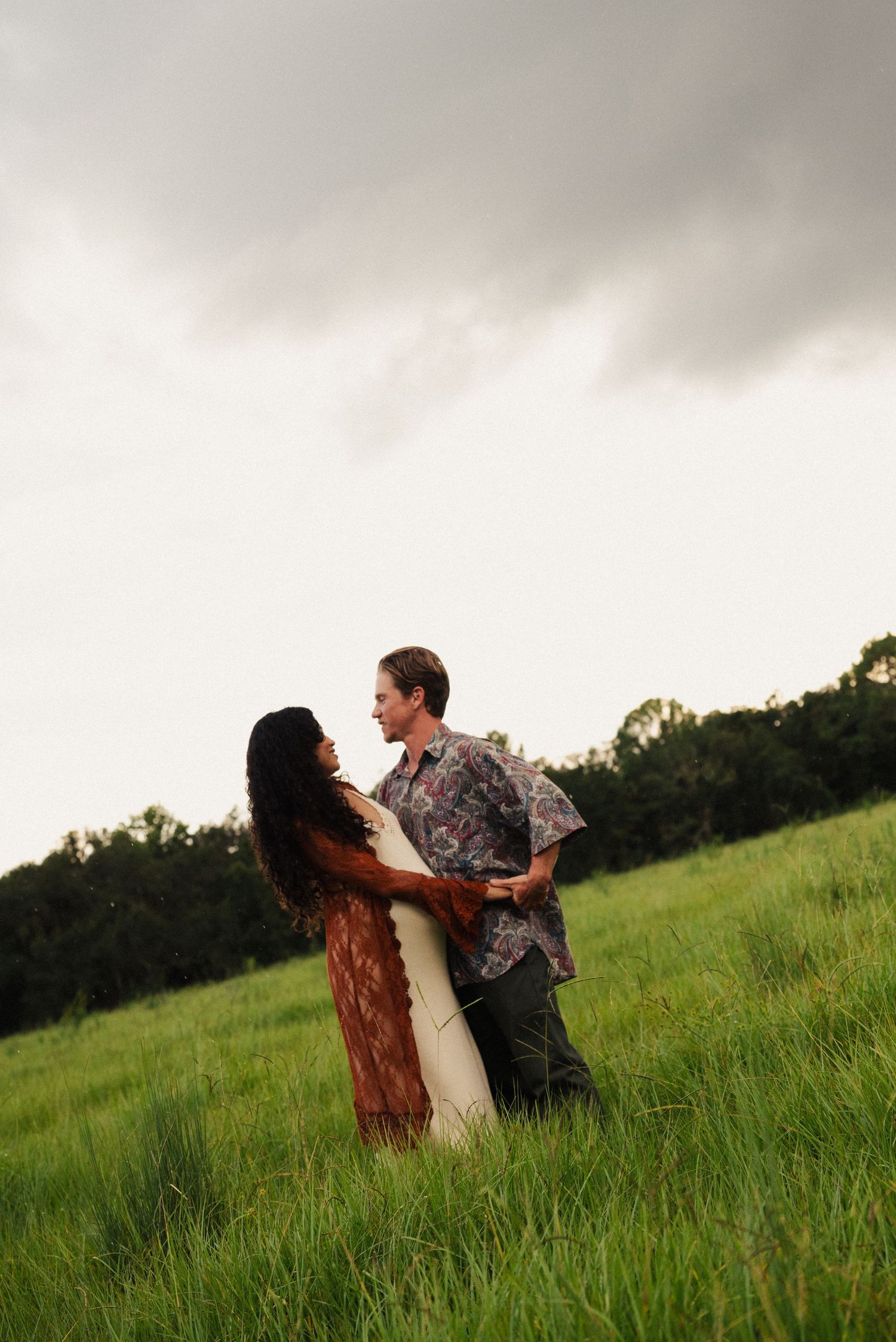 A couple dances together in a dramatic pose in a green field under stormy skies.