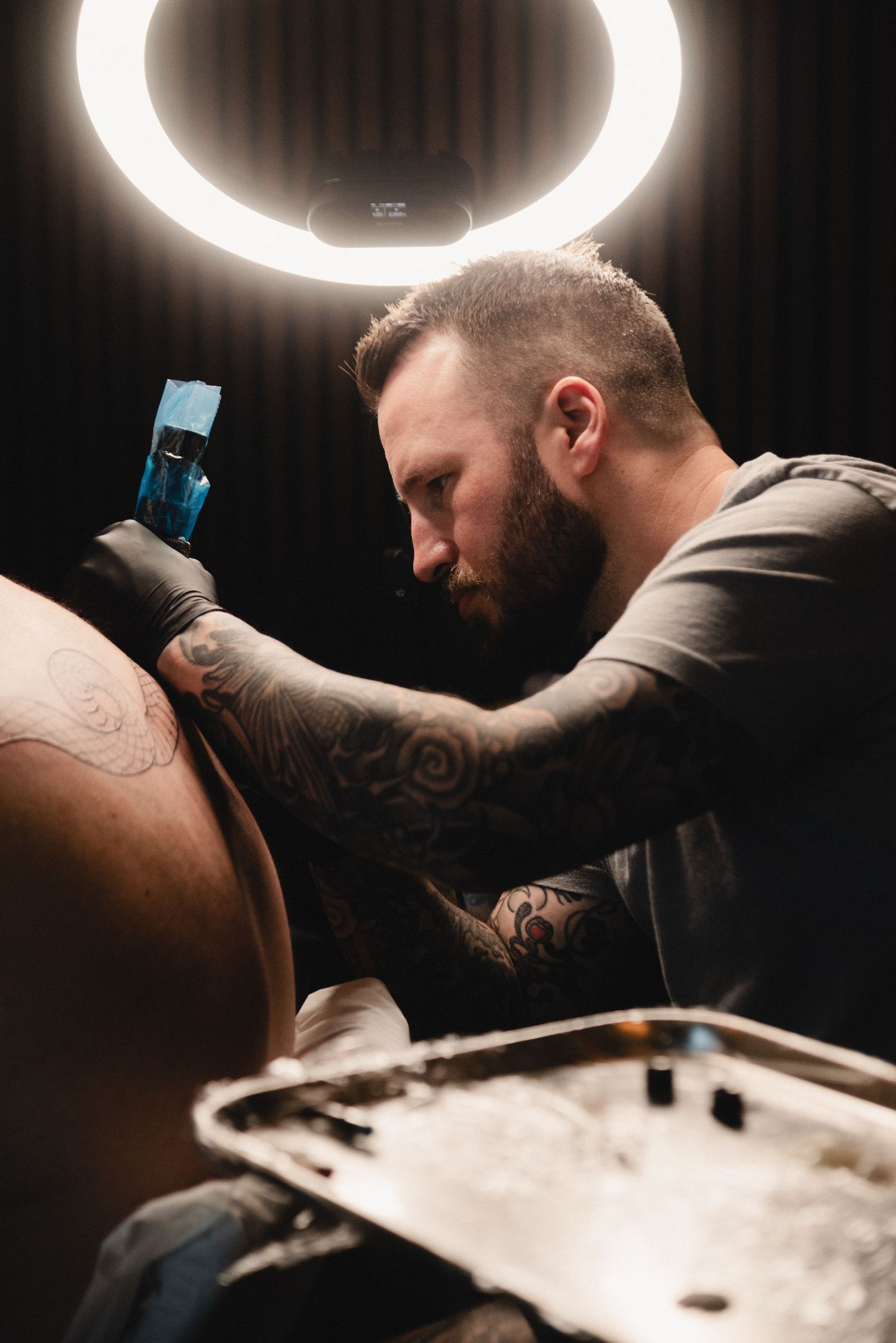 Tattoo artist working under a circular ring light in a professional studio setting.