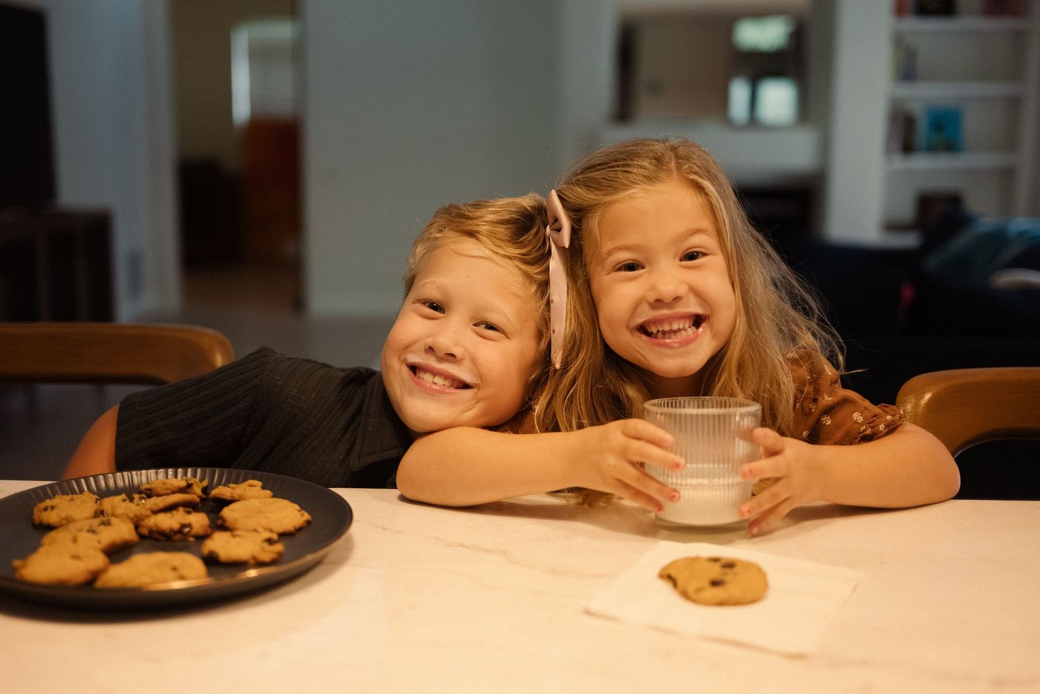 Children smile while leaning on a white counter with cookies visible nearby.