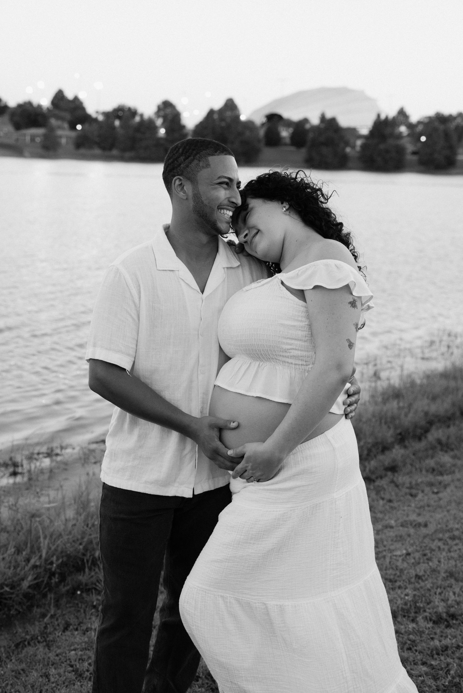 A couple in white outfits embraces lovingly by a lake during a romantic photoshoot.