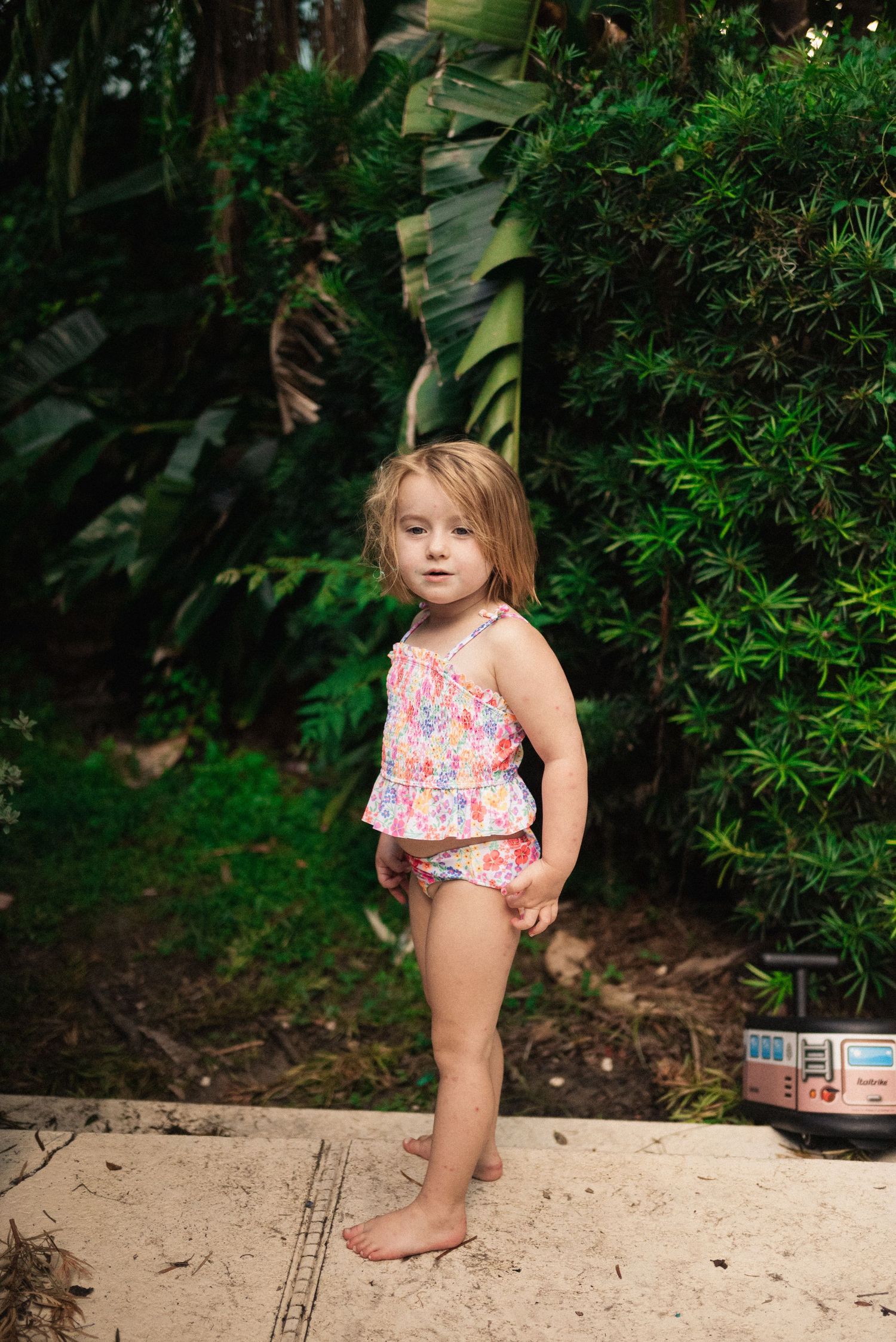A young child in a floral swimsuit plays outdoors near lush greenery.