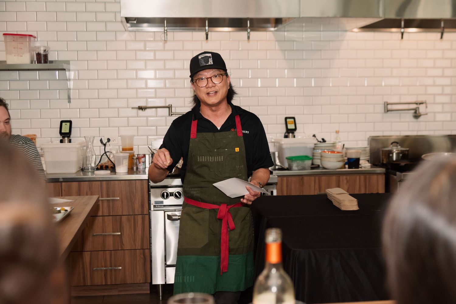 Staff member in green apron and baseball cap working behind wooden counter in restaurant kitchen.