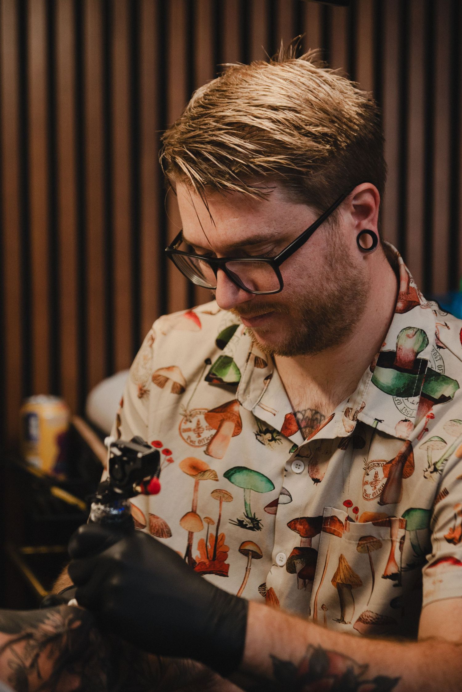 Tattoo artist wearing a colorful mushroom print shirt works intently at their station.