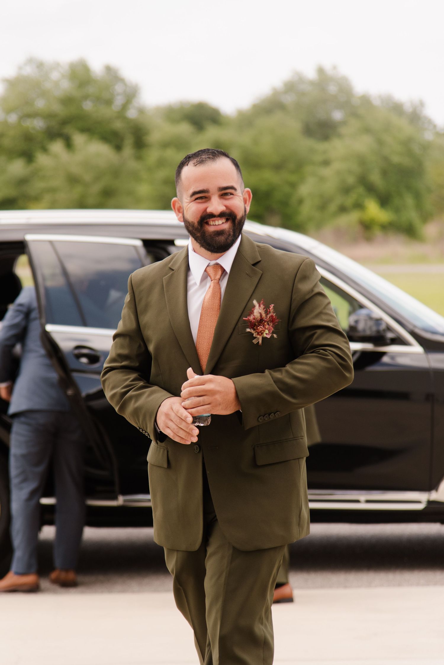 Groomsman in olive suit gives thumbs up near parked cars at outdoor wedding venue.