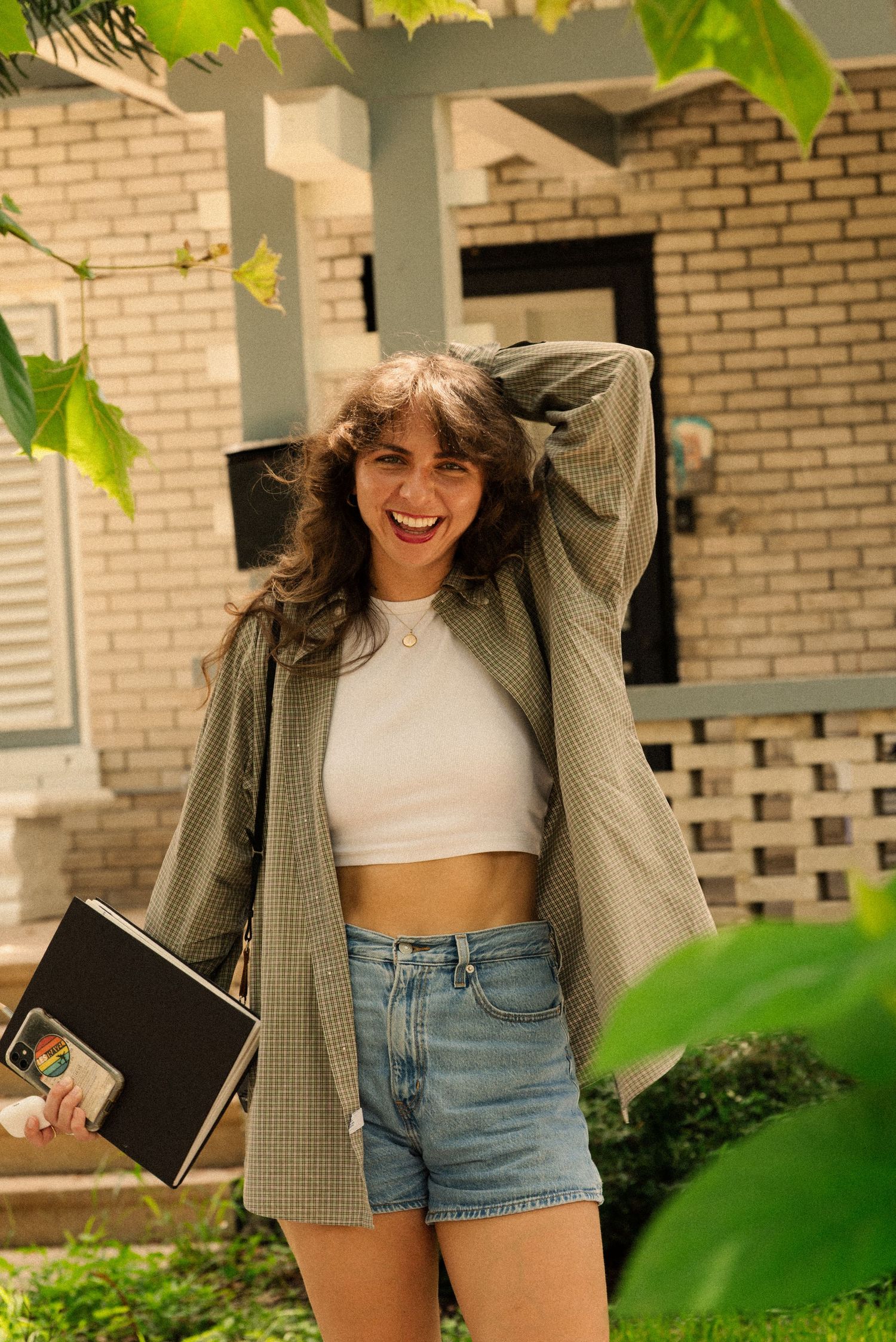 Playful fashion pose in denim shorts and plaid shirt against brick building.