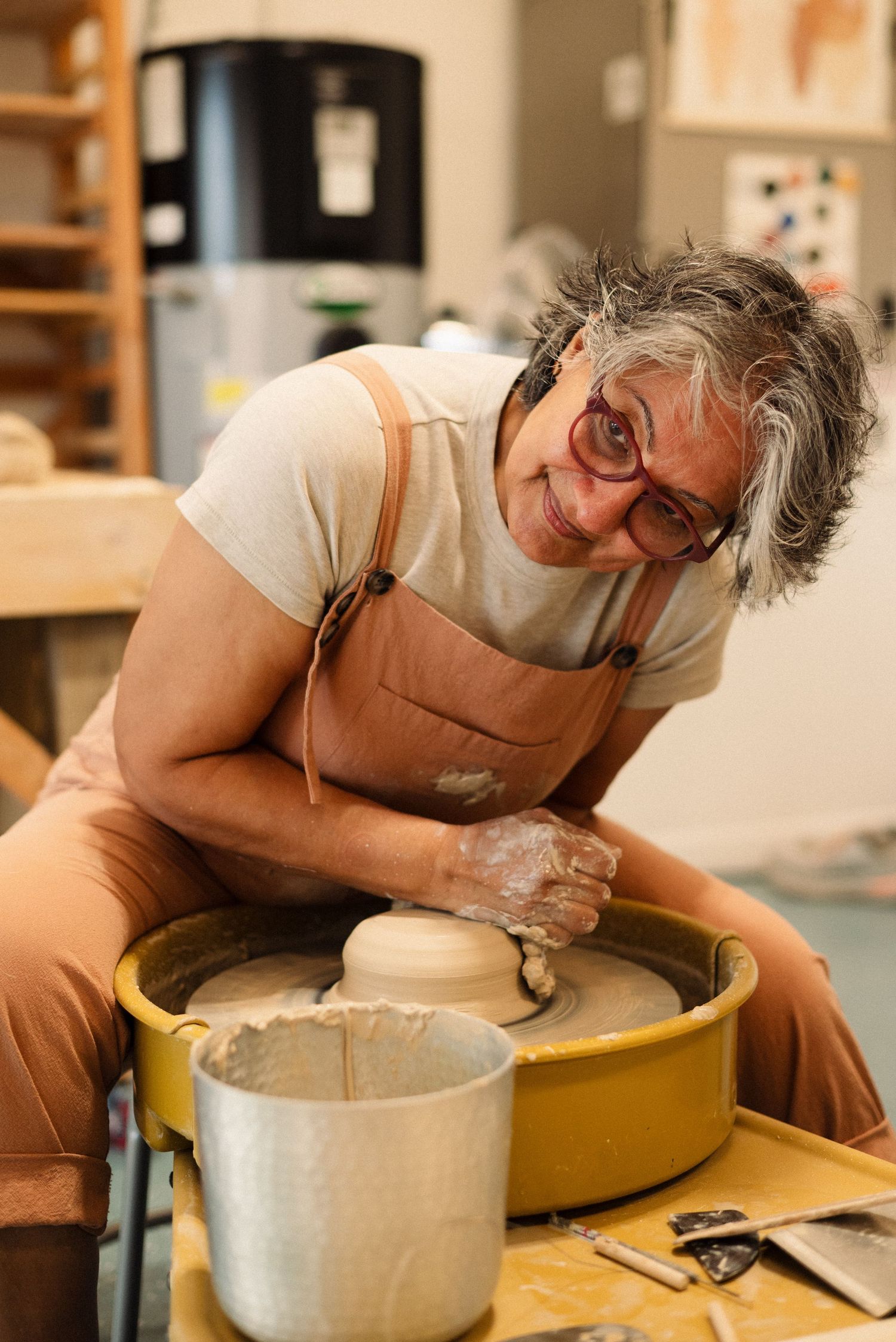 Potter leans over yellow wheel while carefully shaping a ceramic vessel.
