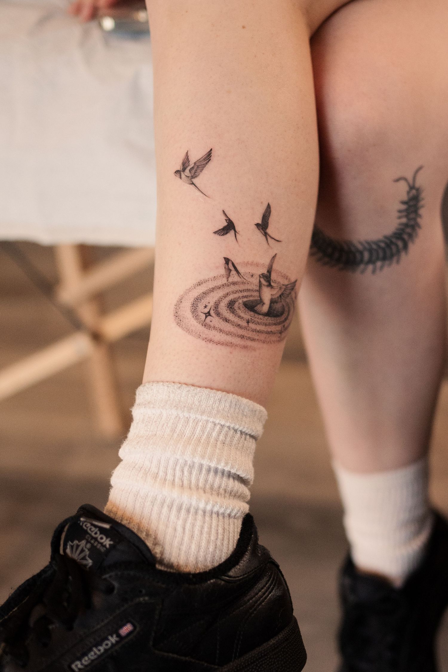 Close-up of delicate bird tattoo designs near white sock and black shoe.
