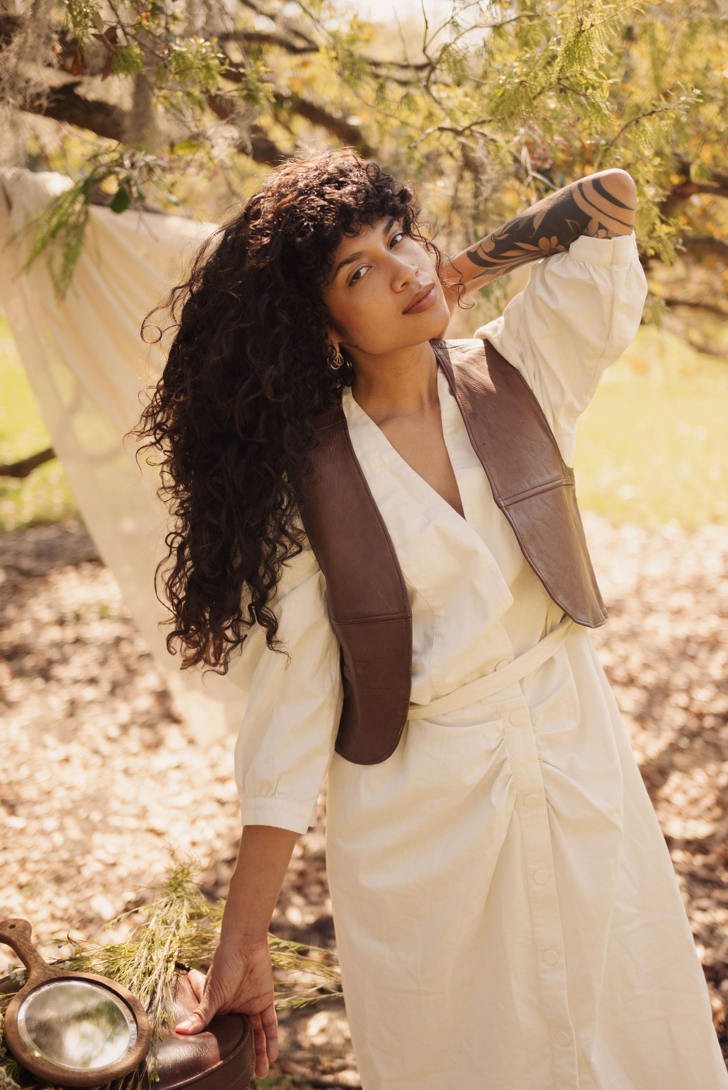 A romantic outdoor fashion portrait featuring a white dress and brown vest in dappled sunlight.